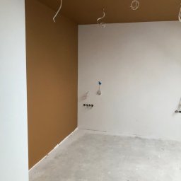 Renovation Hause Wojciech Tworkowski - Pokój w trakcie prac wykończeniowych: pomalowany fragment ściany na brązowo, białe ściany, surowa betonowa podłoga, wystające przewody elektryczne z sufitu i ścian.