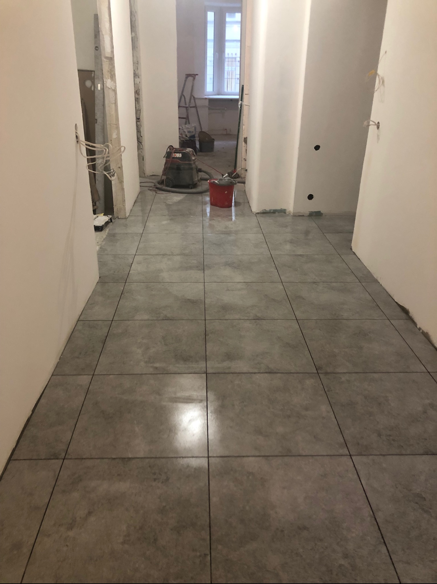 Korytarz w trakcie remontu z ułożonymi szarymi płytkami podłogowymi, widoczne ślady po fugowaniu, odkurzacz przemysłowy i czerwone wiadro w tle, surowe ściany i otwory drzwiowe.