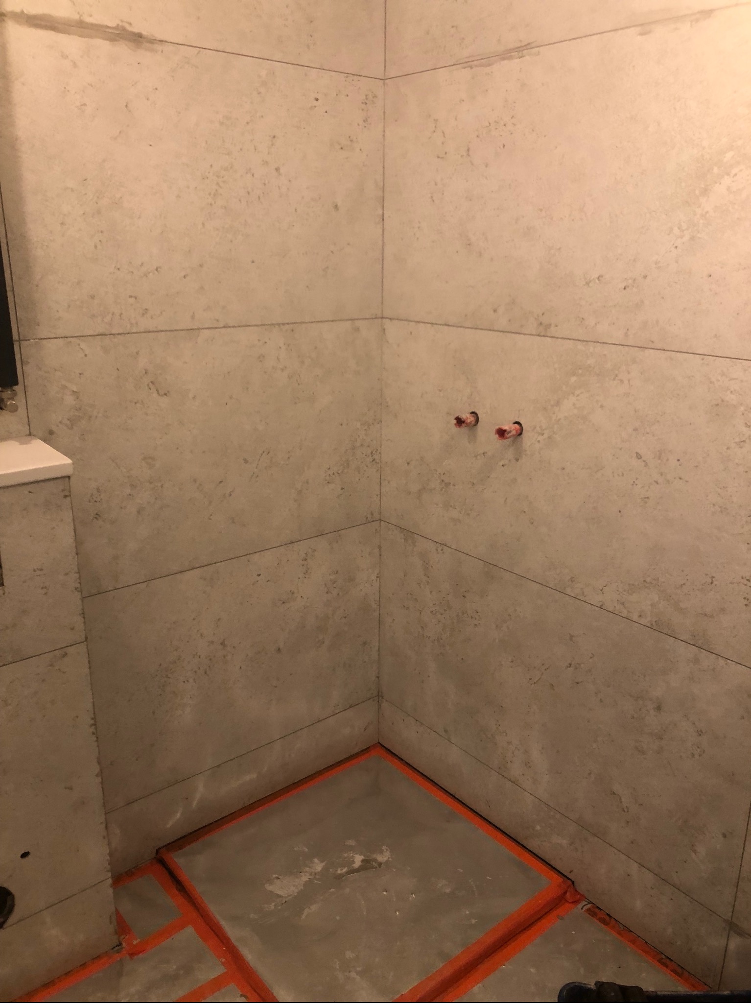 Ściany i podłoga w trakcie kładzenia płytek w łazience, widoczne wystające rury i pomarańczowe listwy oddzielające płytki.