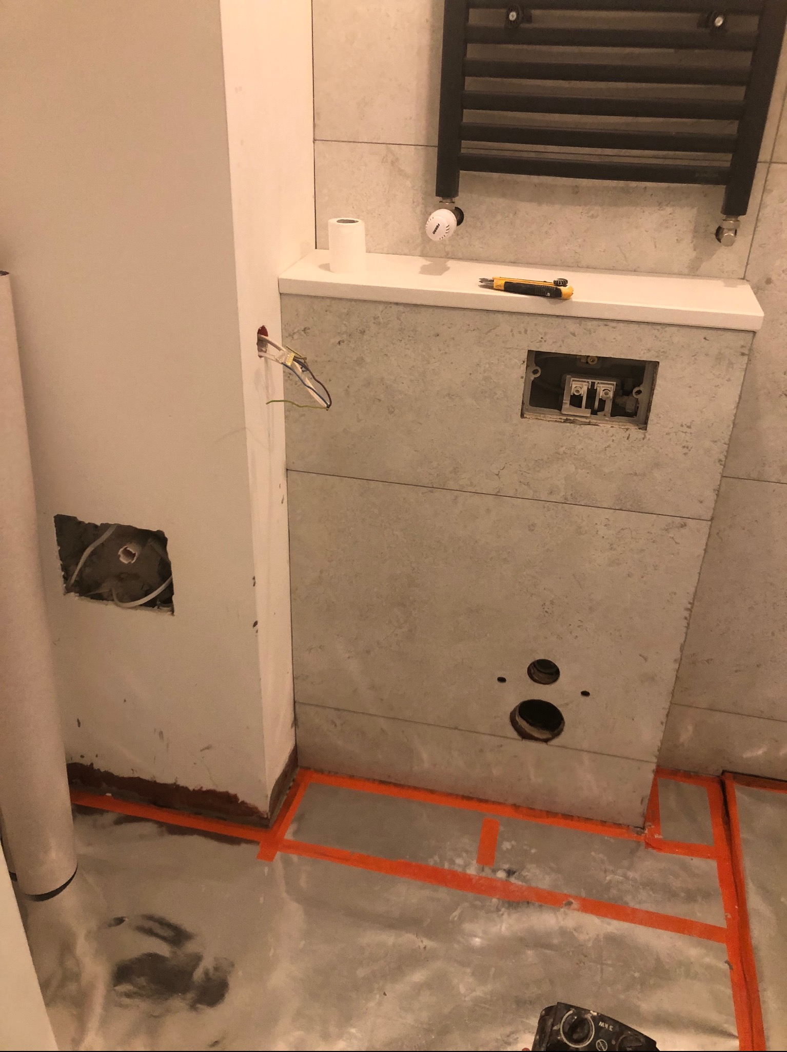 Łazienka w trakcie prac: widoczne przygotowane otwory w ścianie wyłożonej płytkami, wystające przewody elektryczne, zabezpieczona folią podłoga, grzejnik drabinkowy na ścianie i nóż monterski...