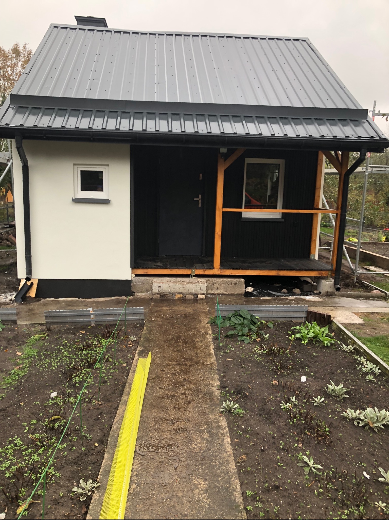 Nowoczesny, parterowy budynek z szarą blachodachówką, czarną elewacją części budynku i białą elewacją innej części, z drewnianym zadaszonym gankiem i elementami rusztowania w tle, otoczony ogrodem...