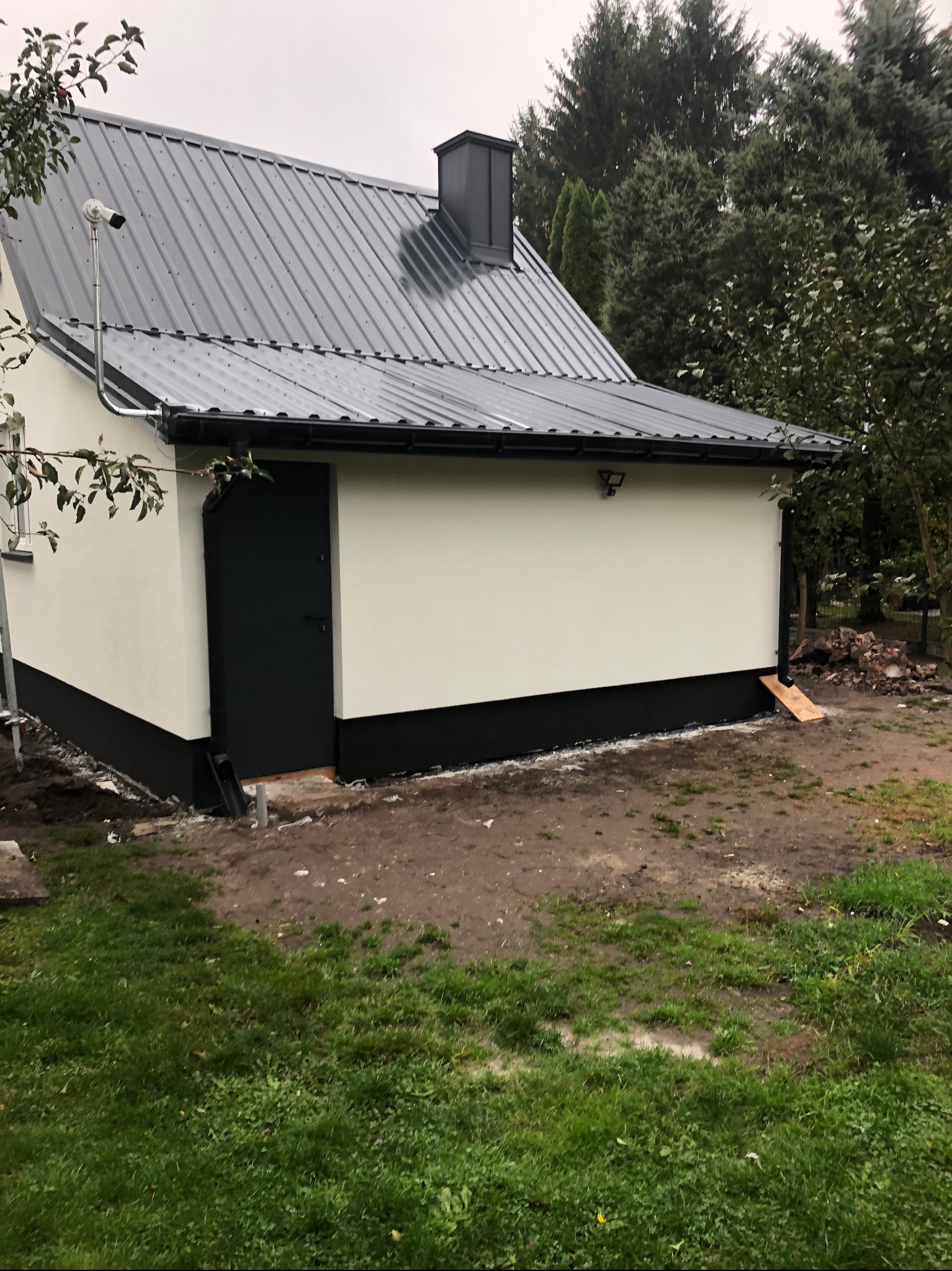 Świeżo otynkowany budynek gospodarczy z grafitową blachodachówką, czarną stolarką i systemem rynnowym, na zewnątrz widoczny teren po budowie i kamera zewnętrzna