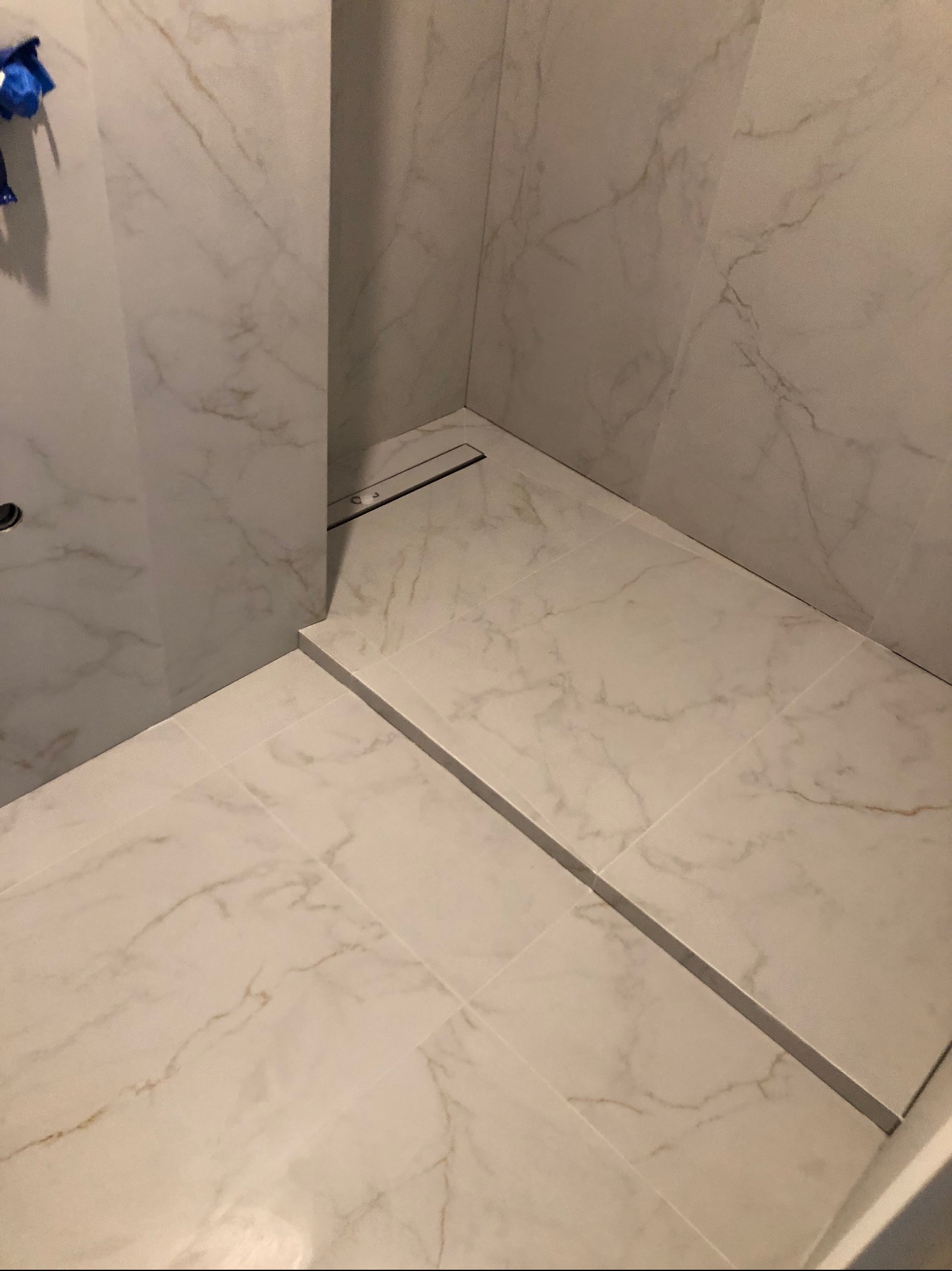 Wykończona łazienka z jasnymi, marmurkowymi płytkami podłogowymi i ściennymi, widoczny liniowy odpływ podłogowy ze stali nierdzewnej.