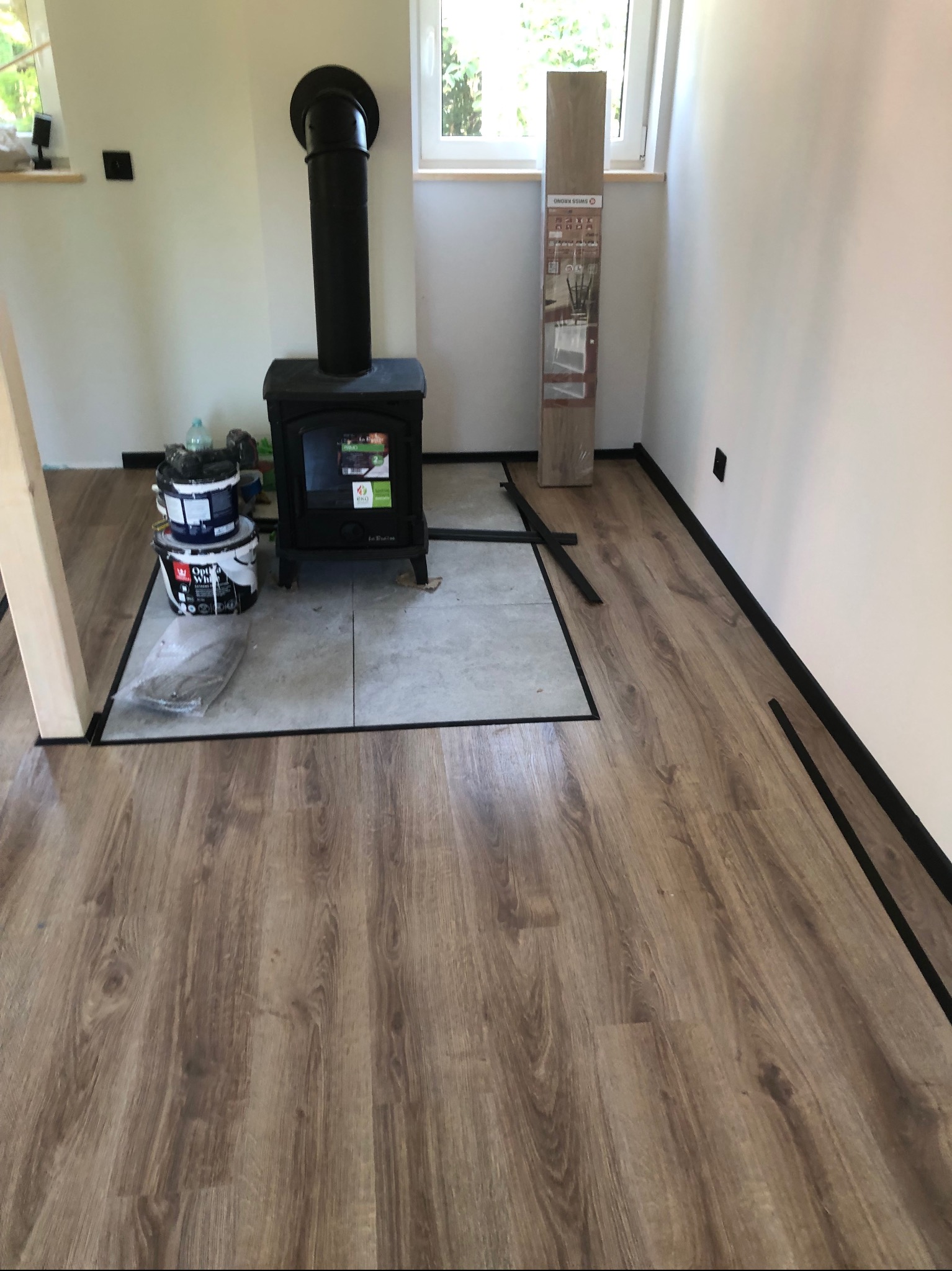 Wnętrze pokoju z nową podłogą z paneli imitujących drewno, czarnym kominkiem na drewno na płytkach, wiaderkami z farbą i listwami przypodłogowymi w trakcie montażu.