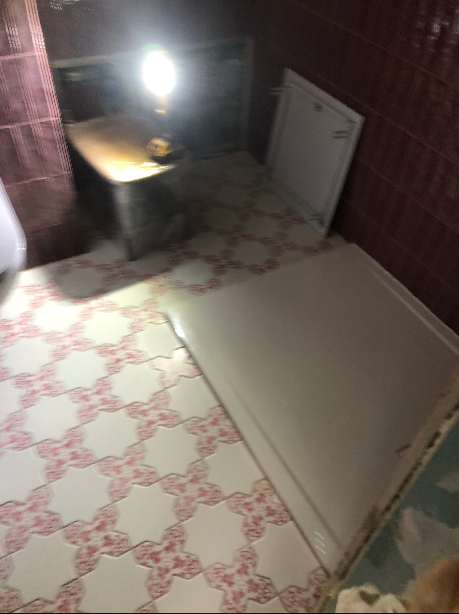 Niedokończona łazienka z białą kabiną prysznicową, wzorzystymi płytkami podłogowymi w odcieniach bieli i różu, oraz fragmentem ściany w bordowych płytkach, oświetlona roboczą lampą.