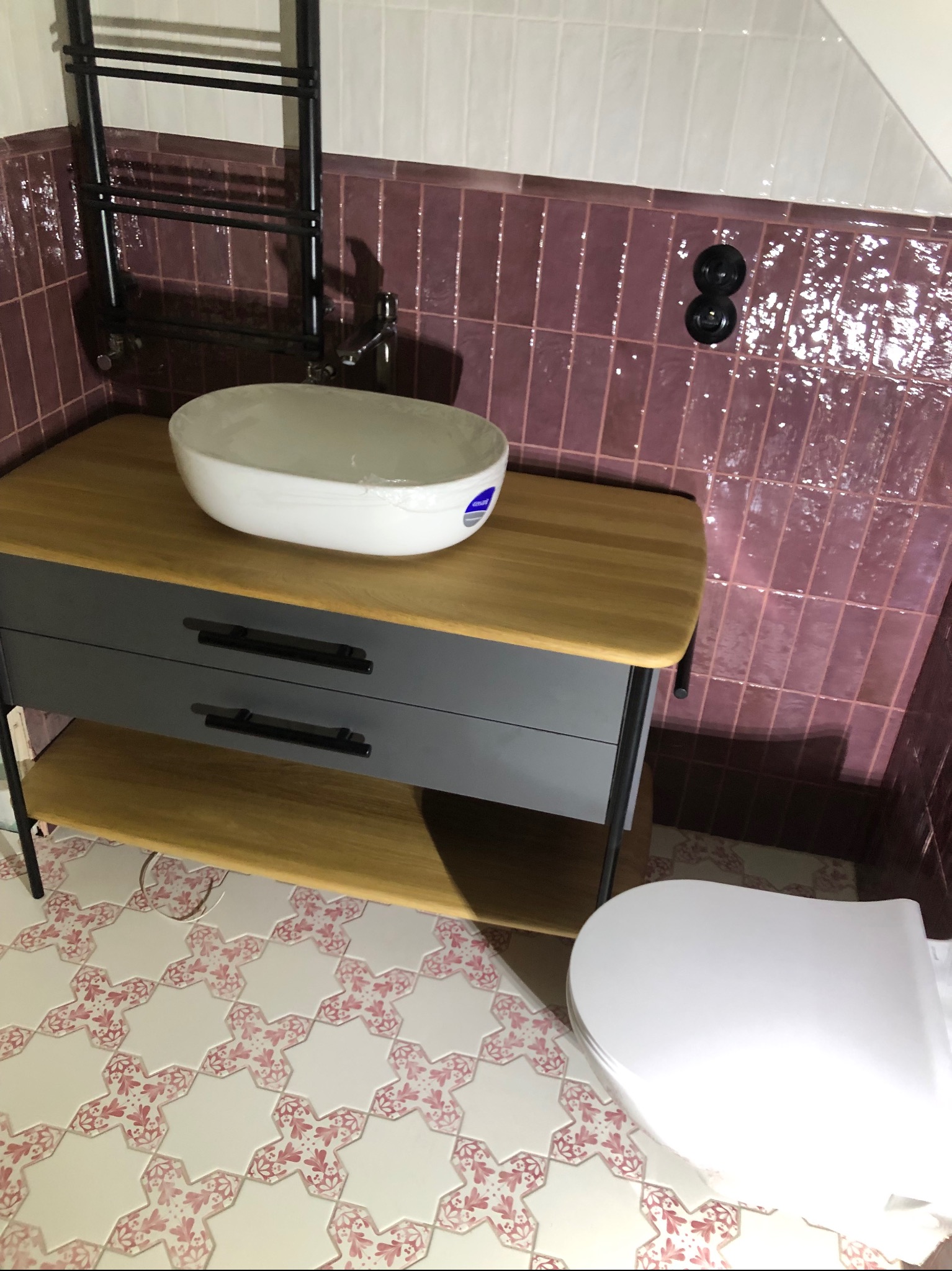 Nowoczesna łazienka z umywalką nablatową na szafce z szufladami, fioletowe płytki na ścianie, ozdobne płytki podłogowe w gwiazdy z różowym wzorem i grzejnik drabinkowy.