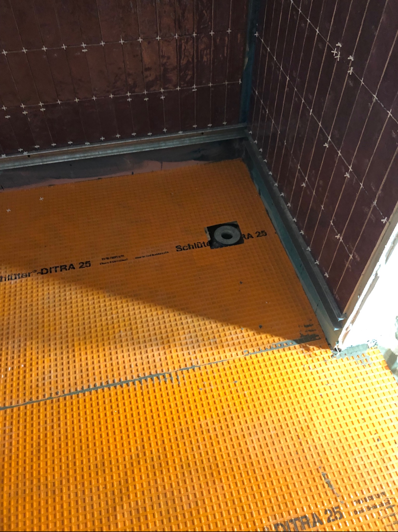 Przygotowanie podłoża pod płytki w kabinie prysznicowej: pomarańczowa mata izolacyjna Schluter-DITRA 25 na podłodze, brązowe płytki ścienne w trakcie układania z krzyżykami dystansowymi.