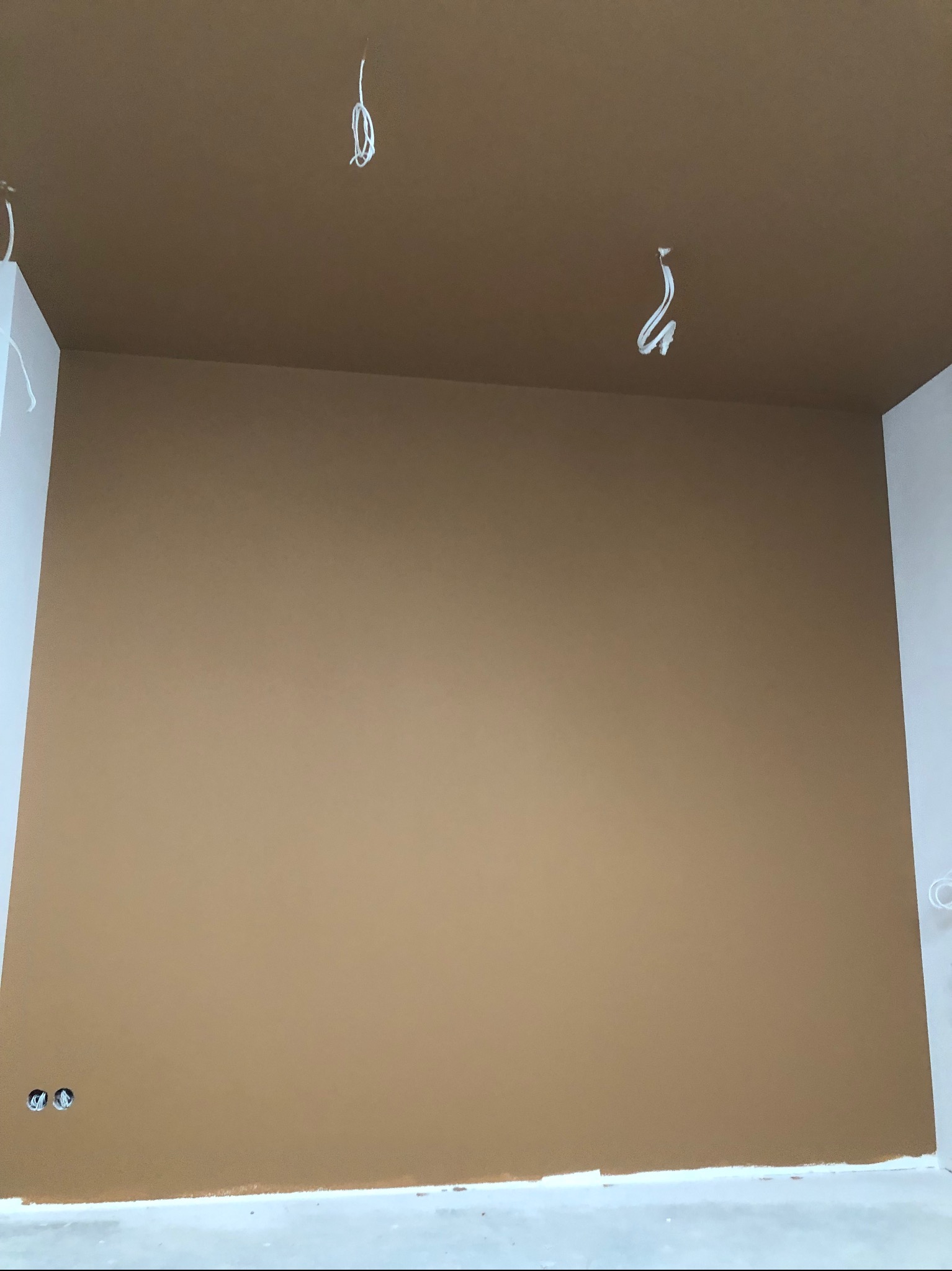 Świeżo pomalowane wnętrze w odcieniach brązu i bieli, widoczne wystające przewody elektryczne z sufitu i gniazdka elektryczne w ścianie, ślady farby na podłodze.