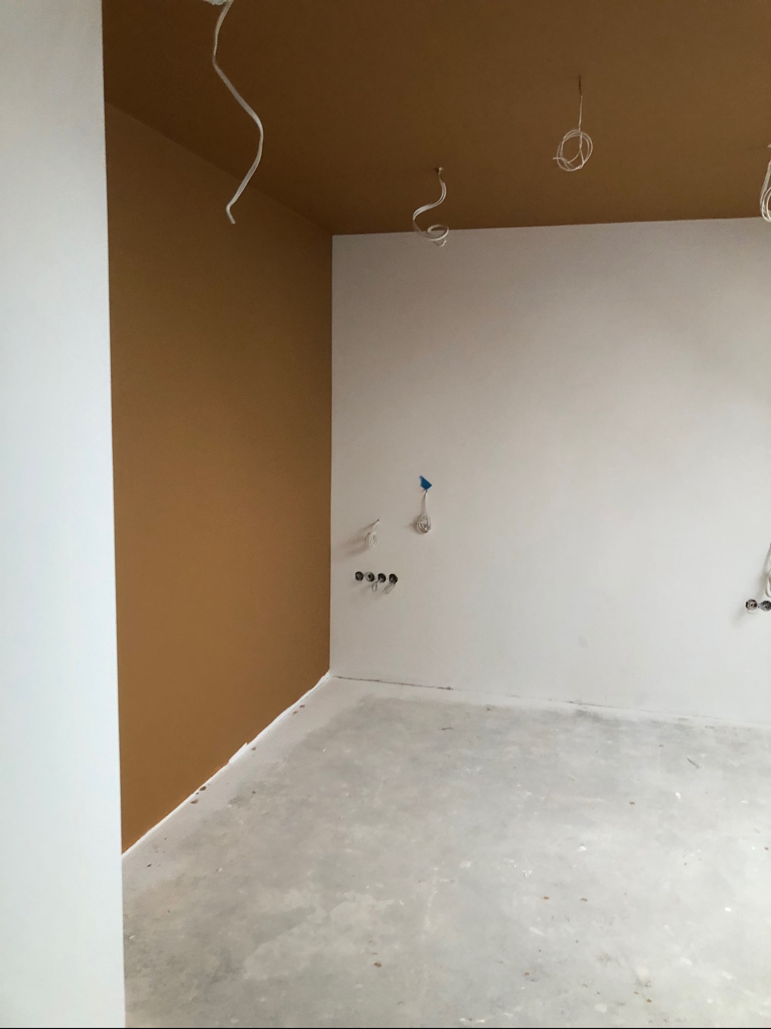 Pokój w trakcie prac wykończeniowych: pomalowany fragment ściany na brązowo, białe ściany, surowa betonowa podłoga, wystające przewody elektryczne z sufitu i ścian.