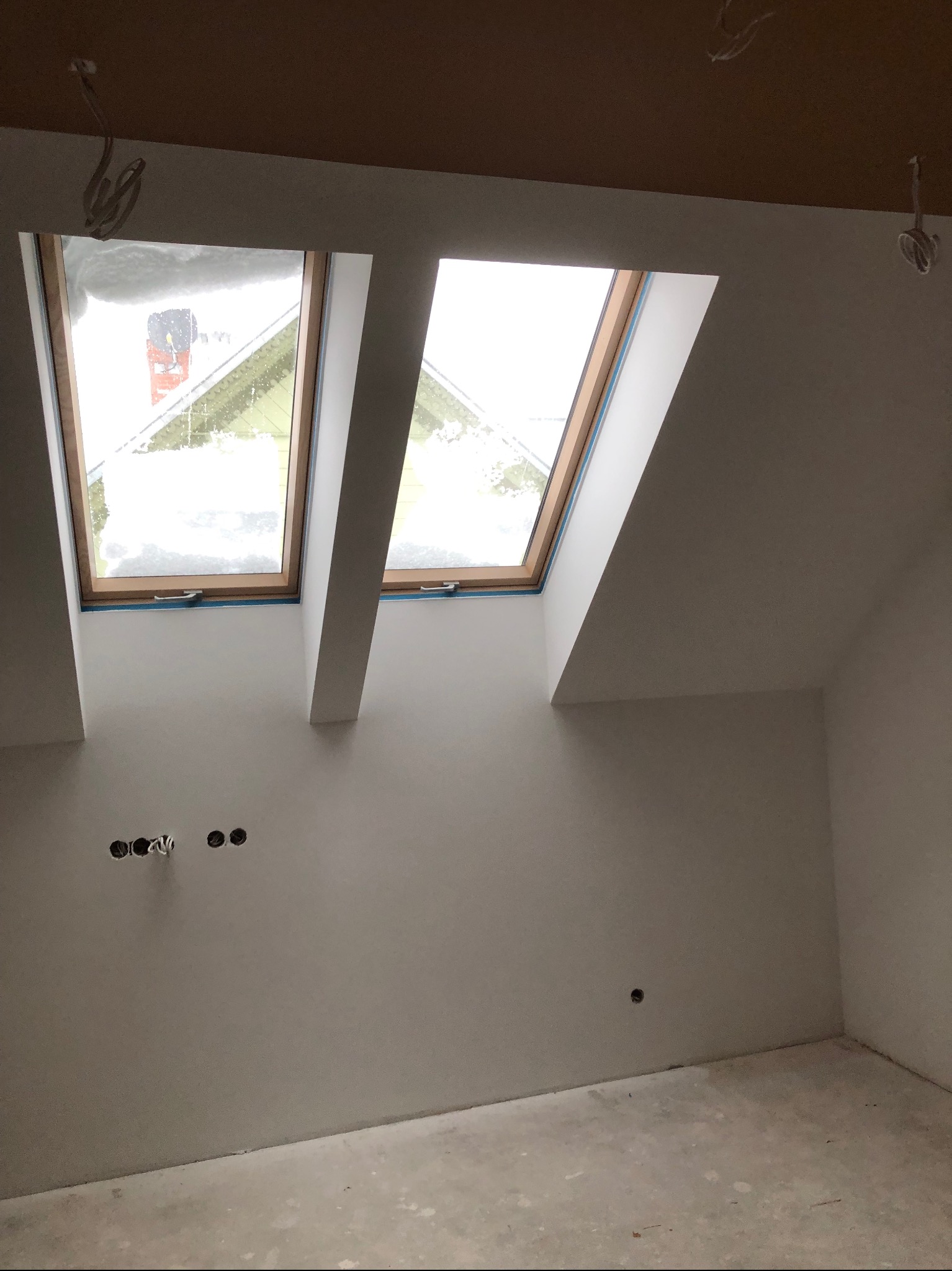 Poddasze z dwoma oknami dachowymi, przez które widać śnieg. Ściany świeżo wyszpachlowane, widoczne puszki elektryczne w ścianie.