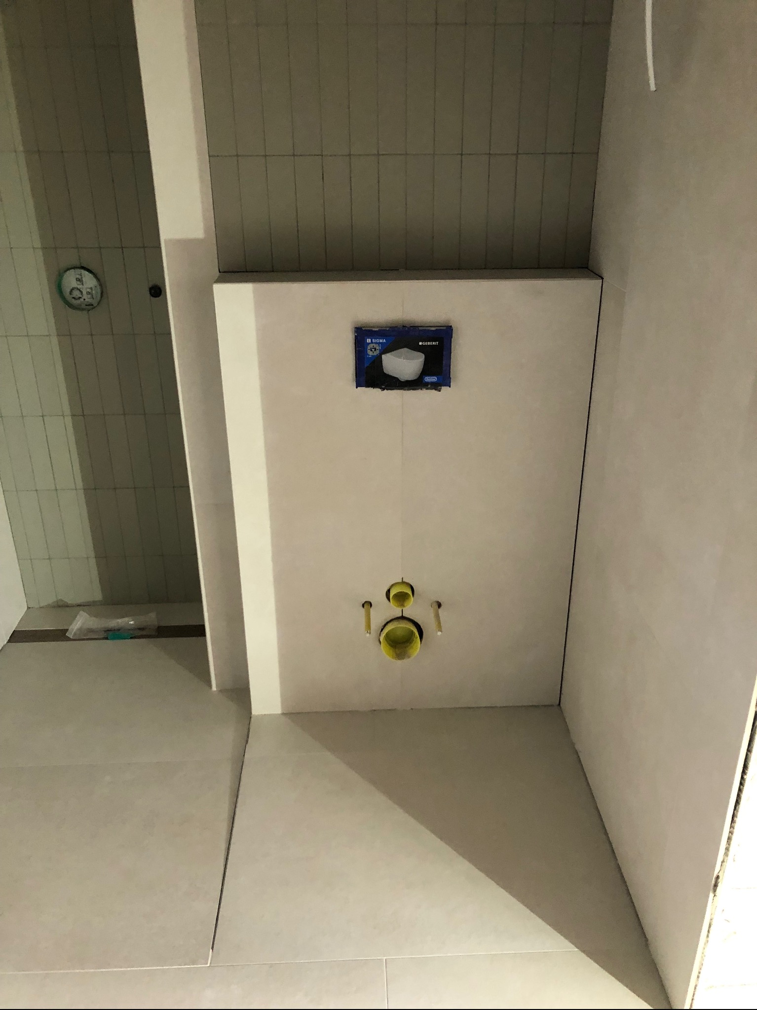 Świeżo wykafelkowana ściana w łazience z zamontowaną podtynkową instalacją WC Geberit, widoczne żółte końcówki zaworów i niebieskie opakowanie na górze.