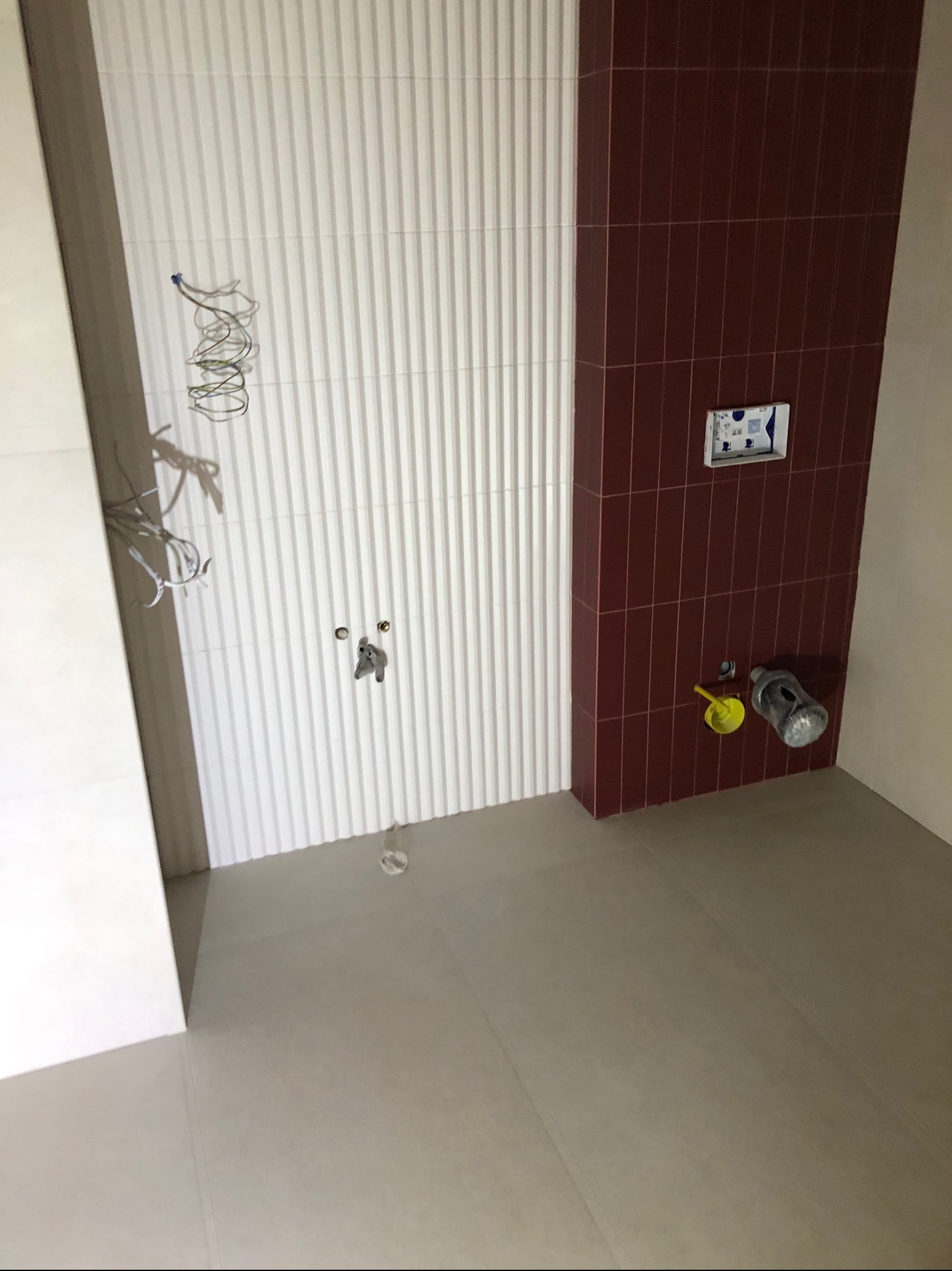 Wnętrze łazienki w trakcie remontu: widoczne pionowe białe panele ścienne, fragment ściany wyłożony wąskimi, bordowymi płytkami, przygotowane przyłącza wodno-kanalizacyjne oraz przewody elektryczne...