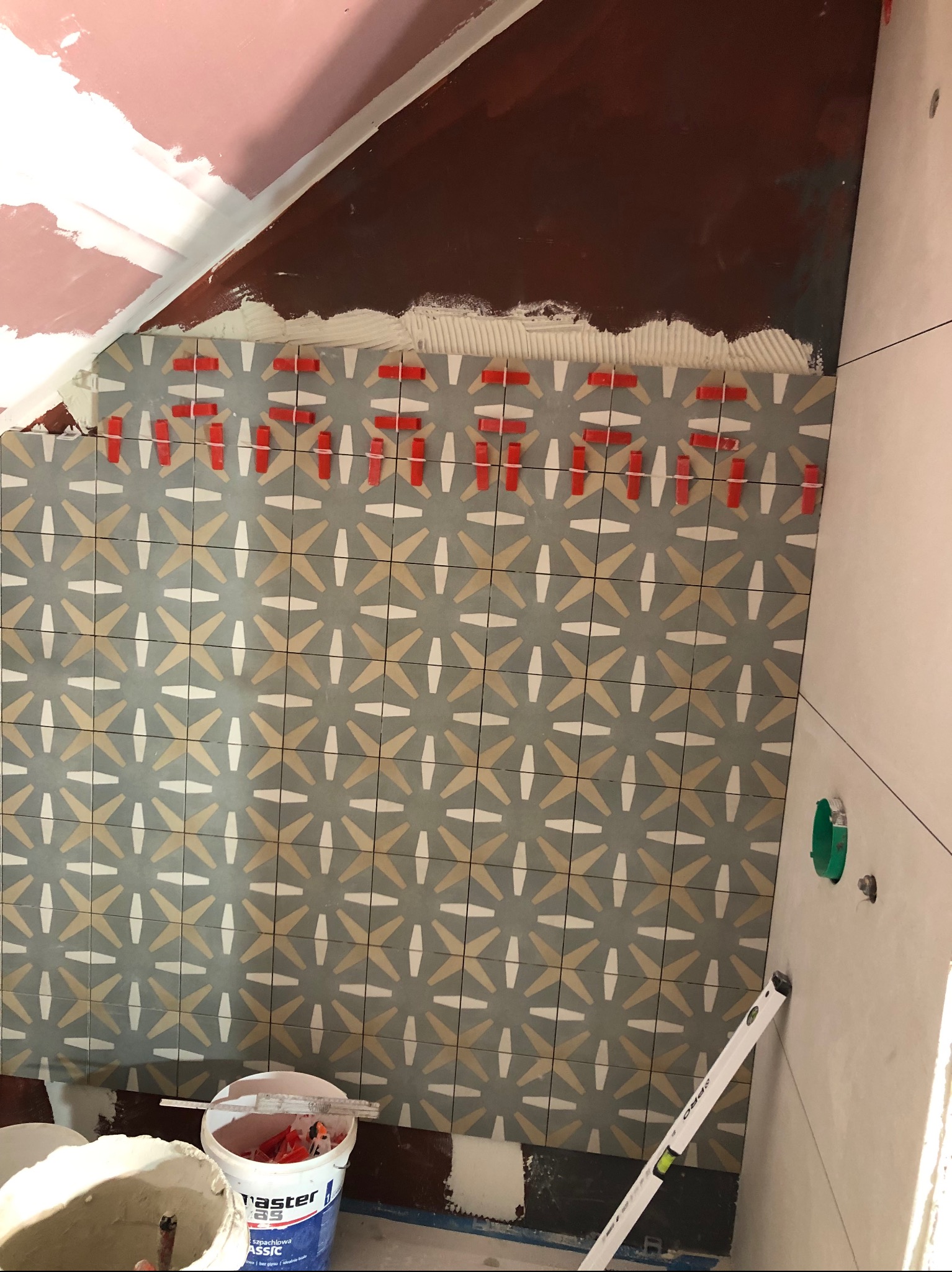 Układanie dekoracyjnych, wzorzystych płytek w łazience z użyciem czerwonych krzyżyków dystansowych, widoczny poziomica i wiaderko z zaprawą szpachlową.