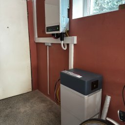 AURUM Energia - Biały inwerter fotowoltaiczny zamontowany na pomarańczowej ścianie obok okna i akumulator energii z logo Pylontech, widoczne przewody i fragment roweru w garażu.