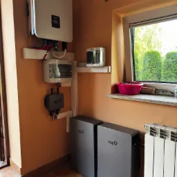 Instalacja domowa: falownik, rozdzielnica i dwa moduły magazynu energii FoxESS zamontowane na pomarańczowej ścianie, widok z dołu, obok okna z miską kasztanów na parapecie.