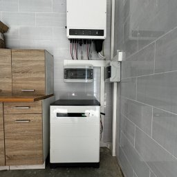 AURUM Energia - Biały magazyn energii Goodwe obok szafki w pomieszczeniu z szarymi płytkami, widoczny inwerter i skrzynka elektryczna.