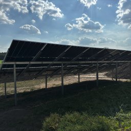 AURUM Energia - Widok od spodu na panele słoneczne zamontowane na metalowej konstrukcji, z widocznym niebem z chmurami w tle, a także fragmentem pola z zielenią i niewielkim pagórkiem ziemi.