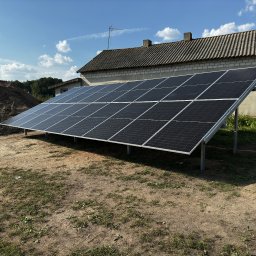 AURUM Energia - Instalacja paneli słonecznych na gruncie przed budynkiem z szarej cegły i stertą ziemi w tle, błękitne niebo z chmurami.