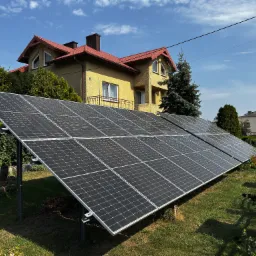 Panele fotowoltaiczne na trawniku przed domem z czerwoną dachówką, widok z niskiej perspektywy, słoneczny dzień.