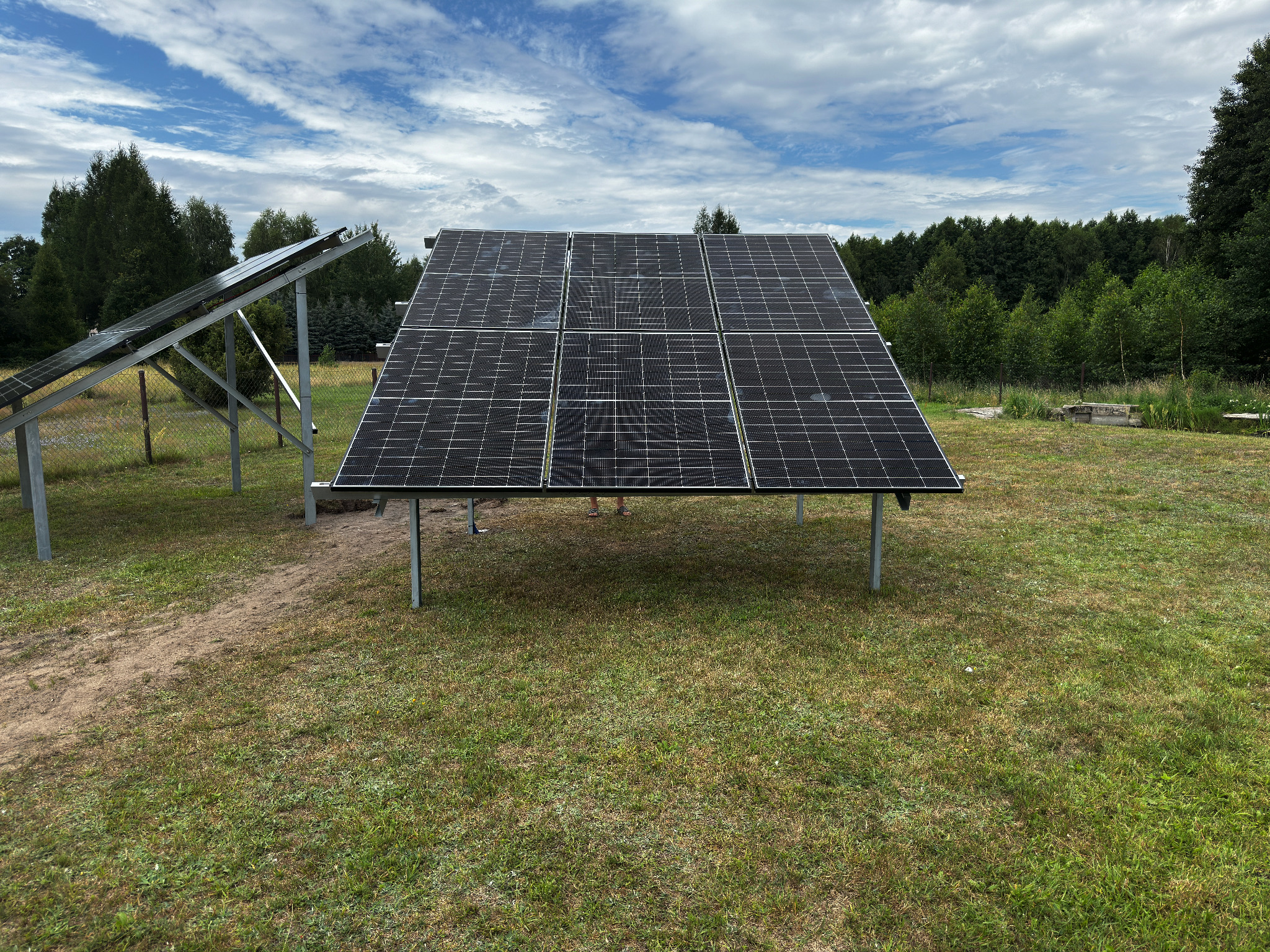 Instalacja fotowoltaiczna na trawiastym terenie z widocznymi drzewami w tle i błękitnym niebem z chmurami. Pod modułami widoczne stopy osoby.