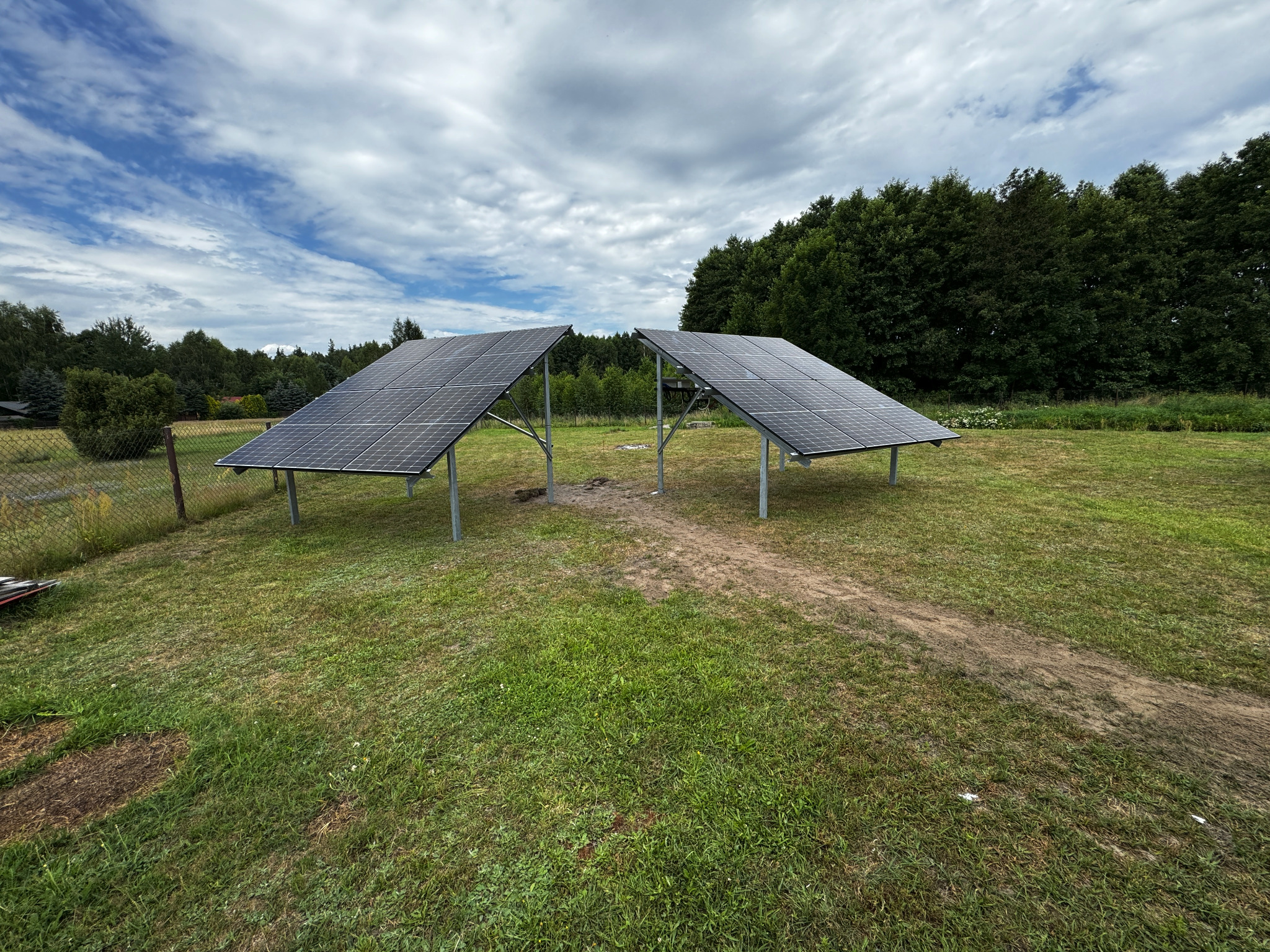 Dwa panele słoneczne ustawione na metalowych stelażach na trawiastym terenie, z widocznym śladem przejazdu między nimi, w tle drzewa i pochmurne niebo.