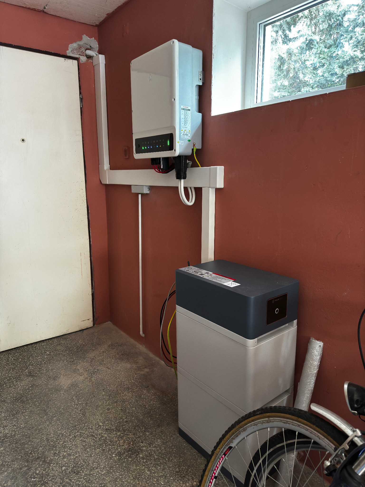 Biały inwerter fotowoltaiczny zamontowany na pomarańczowej ścianie obok okna i akumulator energii z logo Pylontech, widoczne przewody i fragment roweru w garażu.