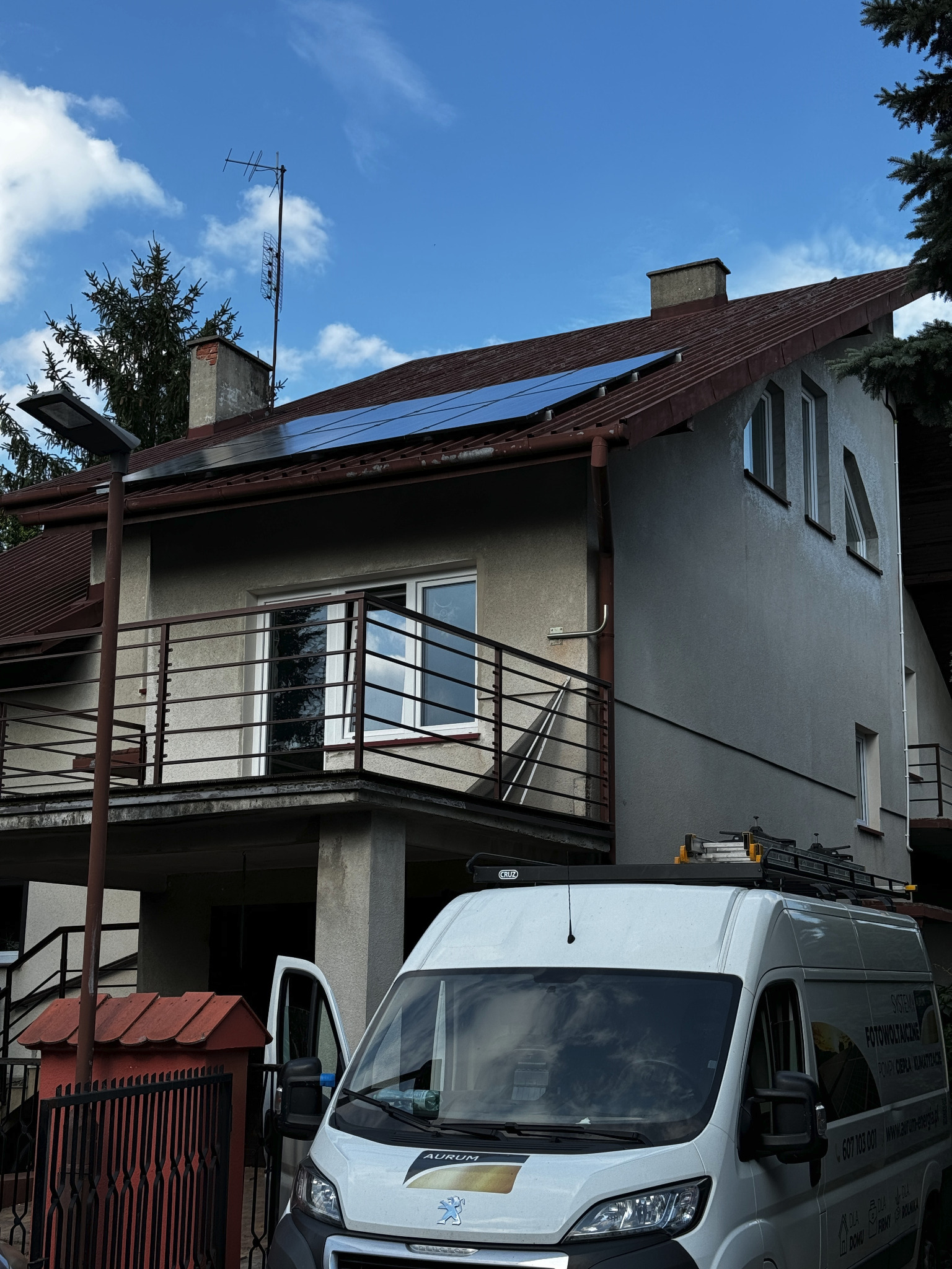 Montaż paneli fotowoltaicznych na dachu dwupiętrowego domu z szarą elewacją, widoczny samochód dostawczy z logo firmy Aurum oraz numerem telefonu, drabina na dachu samochodu