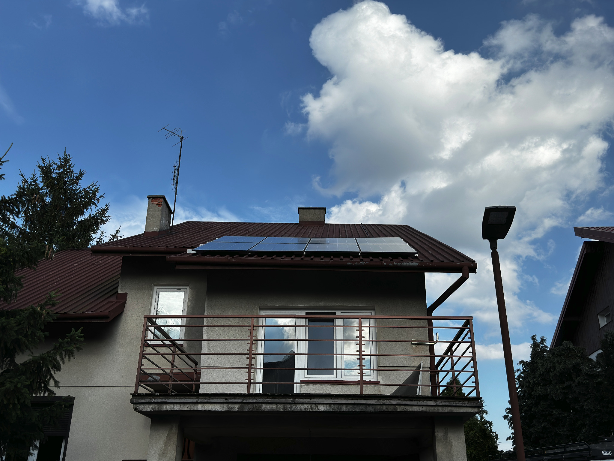 Dom jednorodzinny z zamontowanymi panelami słonecznymi na dachu z blachodachówki w pochmurny dzień. Na balkonie widoczne krzesła.