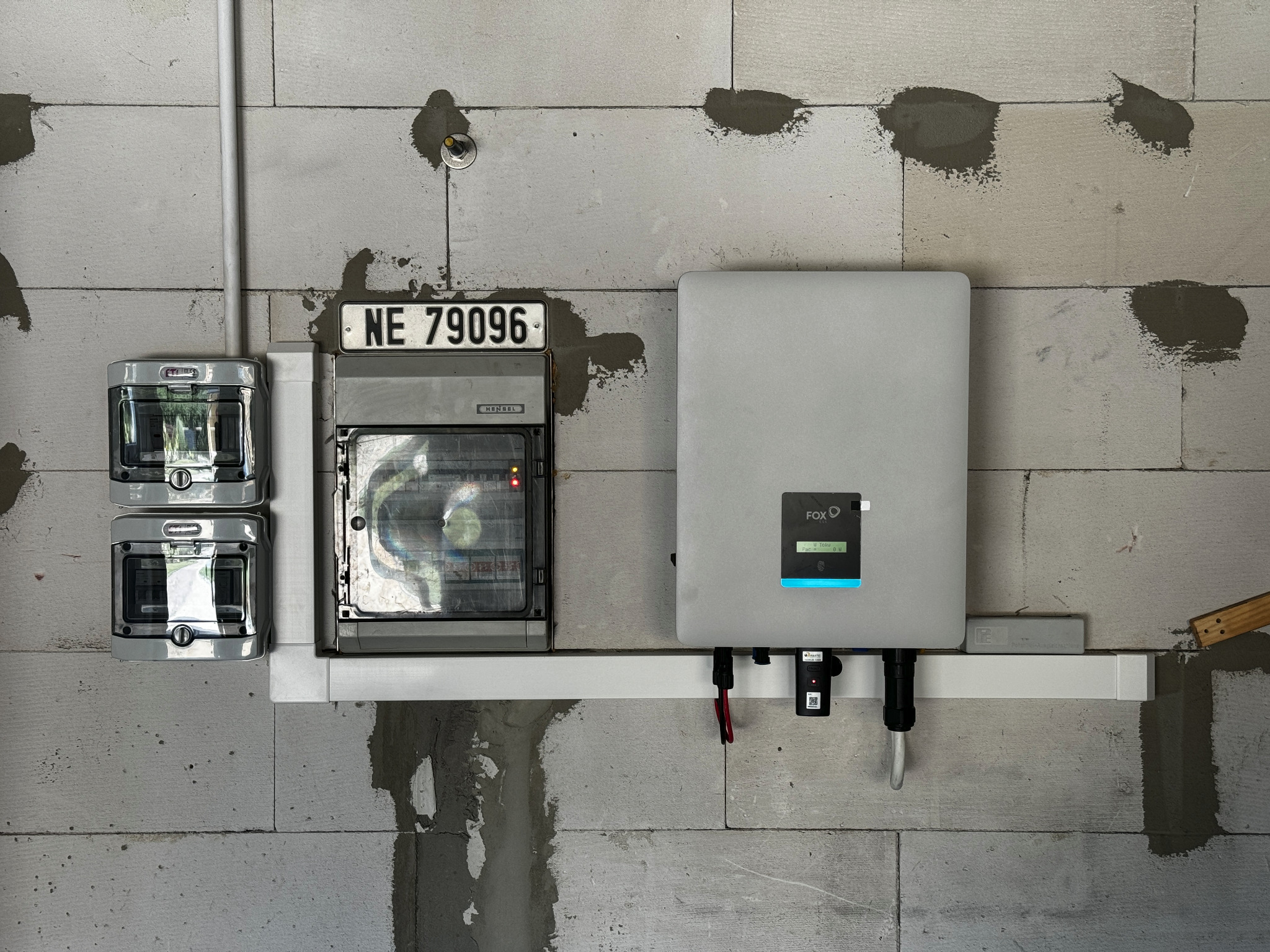Szary inwerter marki FOX-ESS z podłączonymi kablami, zamontowany na białej listwie przy ścianie z betonu komórkowego z widocznymi śladami po naprawach, obok skrzynka elektryczna z numerem NE 79096.