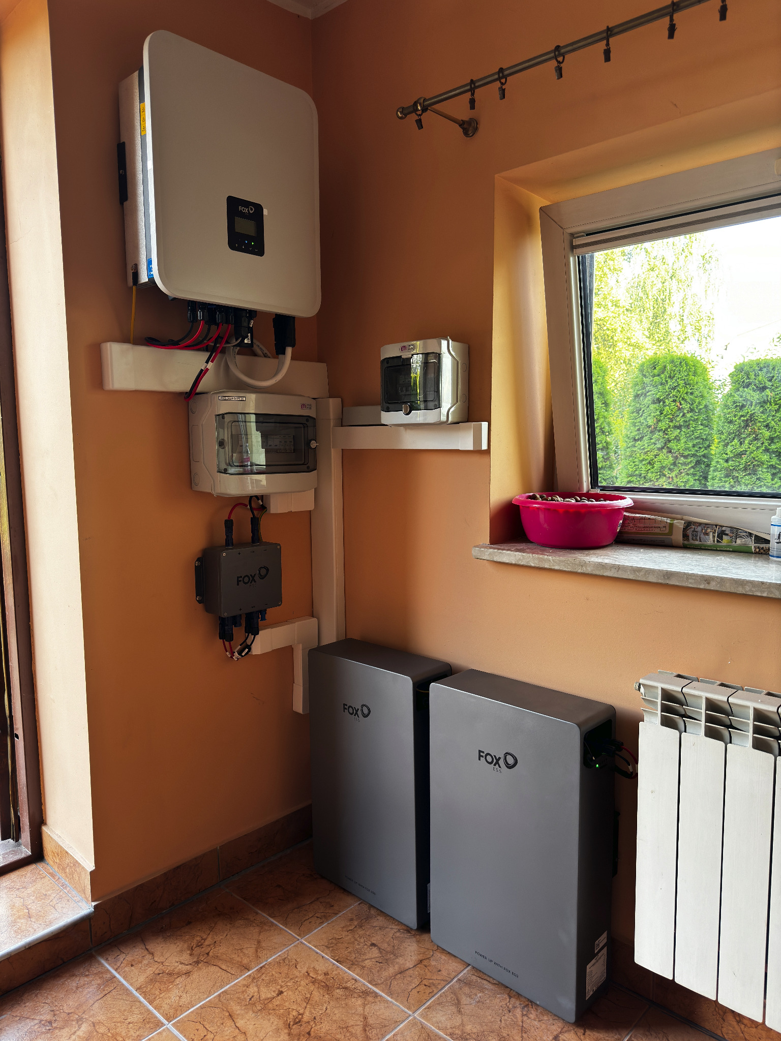 Instalacja domowa: falownik, rozdzielnica i dwa moduły magazynu energii FoxESS zamontowane na pomarańczowej ścianie, widok z dołu, obok okna z miską kasztanów na parapecie.