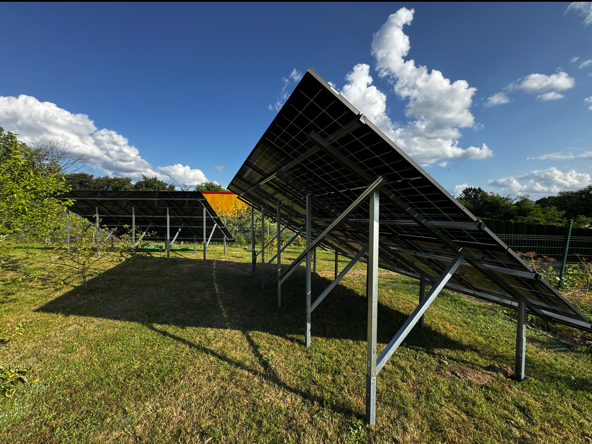 Panele słoneczne ustawione na metalowych stelażach na trawiastym terenie w słoneczny dzień z błękitnym niebem i białymi chmurami w tle.