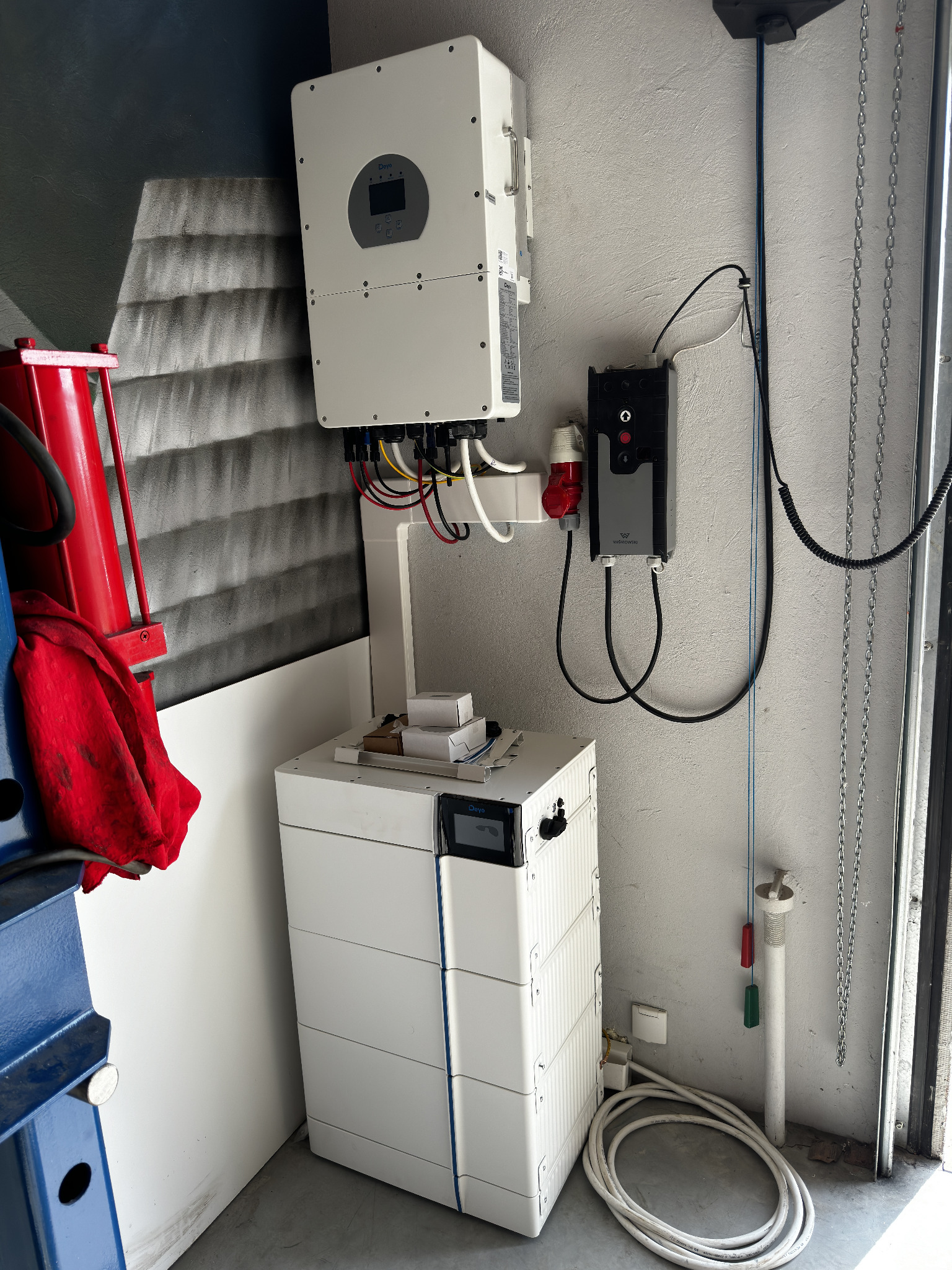 Biały inwerter fotowoltaiczny marki Deye zamontowany na ścianie obok modułowej baterii magazynu energii z widocznymi kablami i elementami instalacji elektrycznej w pomieszczeniu gospodarczym.