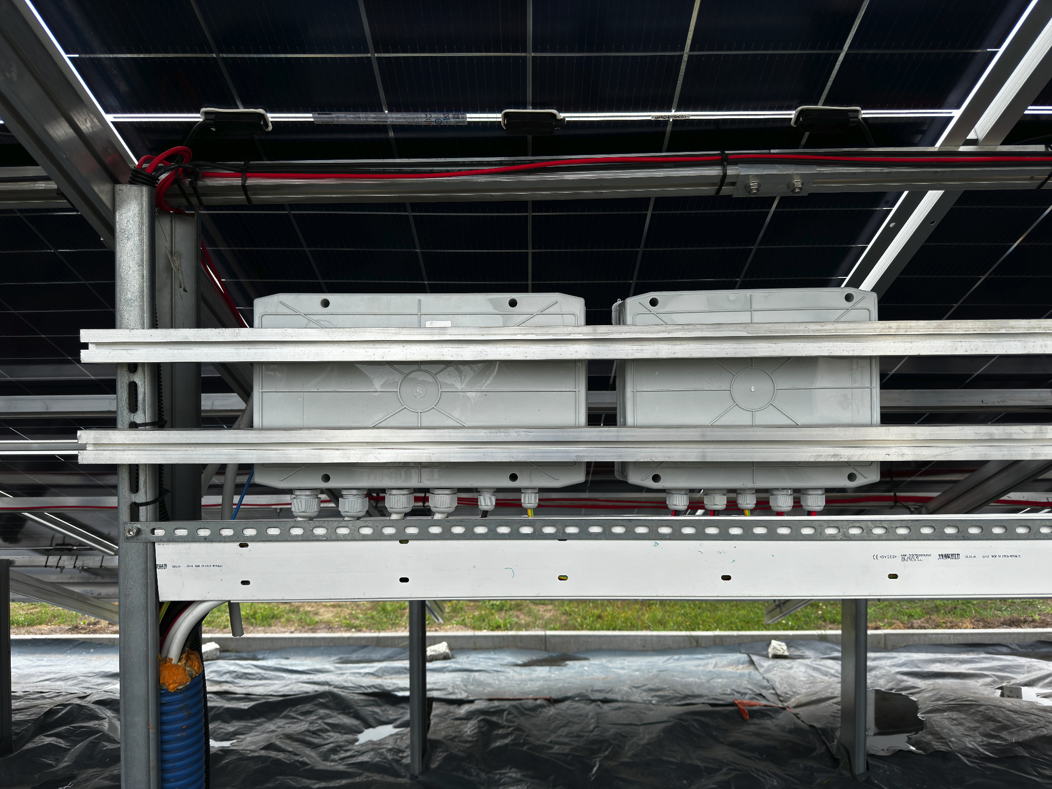 Widok od spodu na konstrukcję wsporczą paneli słonecznych z widocznymi puszkami przyłączeniowymi, przewodami i elementami montażowymi, z zielenią w tle.