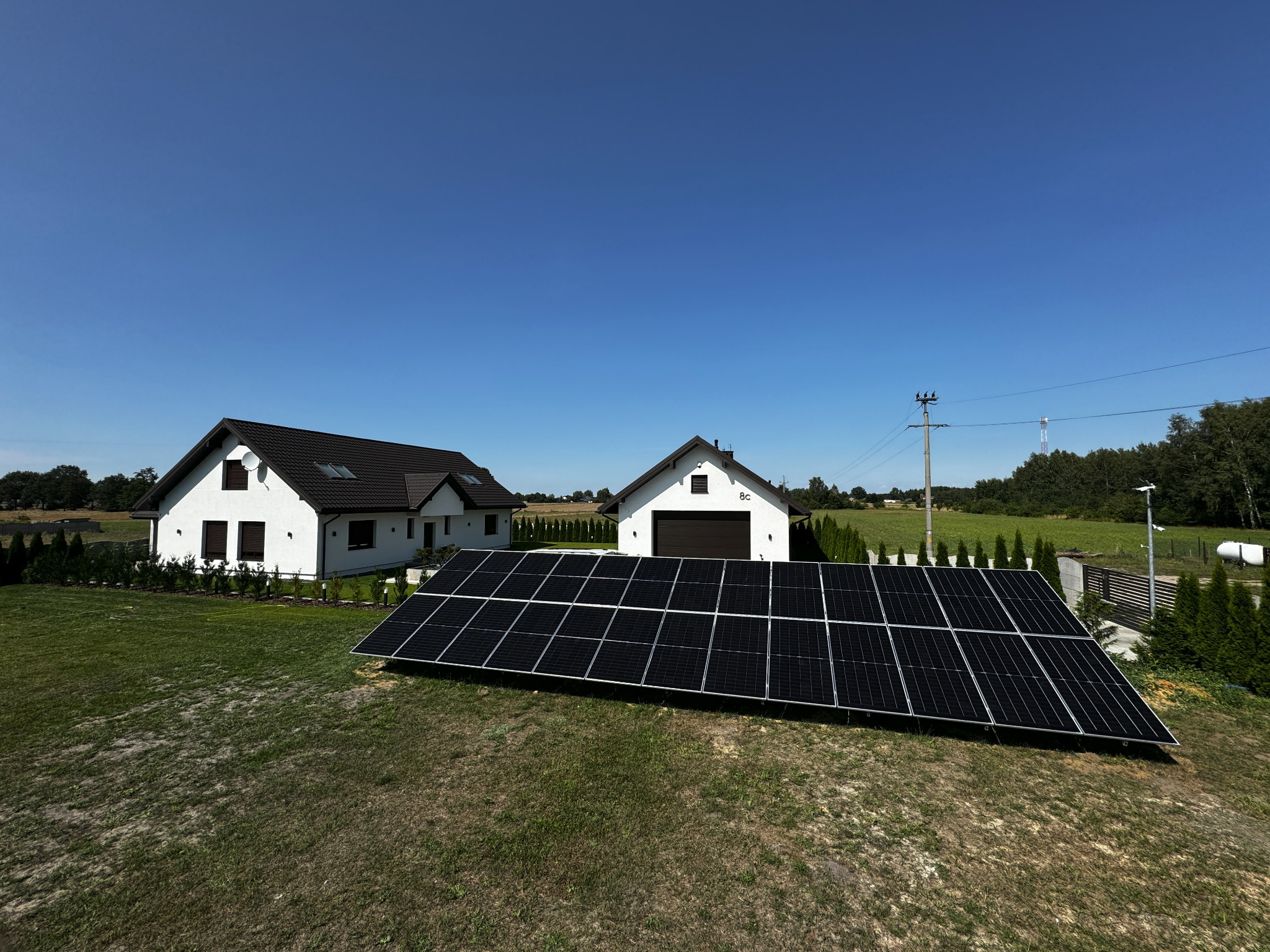 Panele słoneczne na trawniku przed nowoczesnym domem z białymi ścianami i brązowym dachem, błękitne niebo w tle.