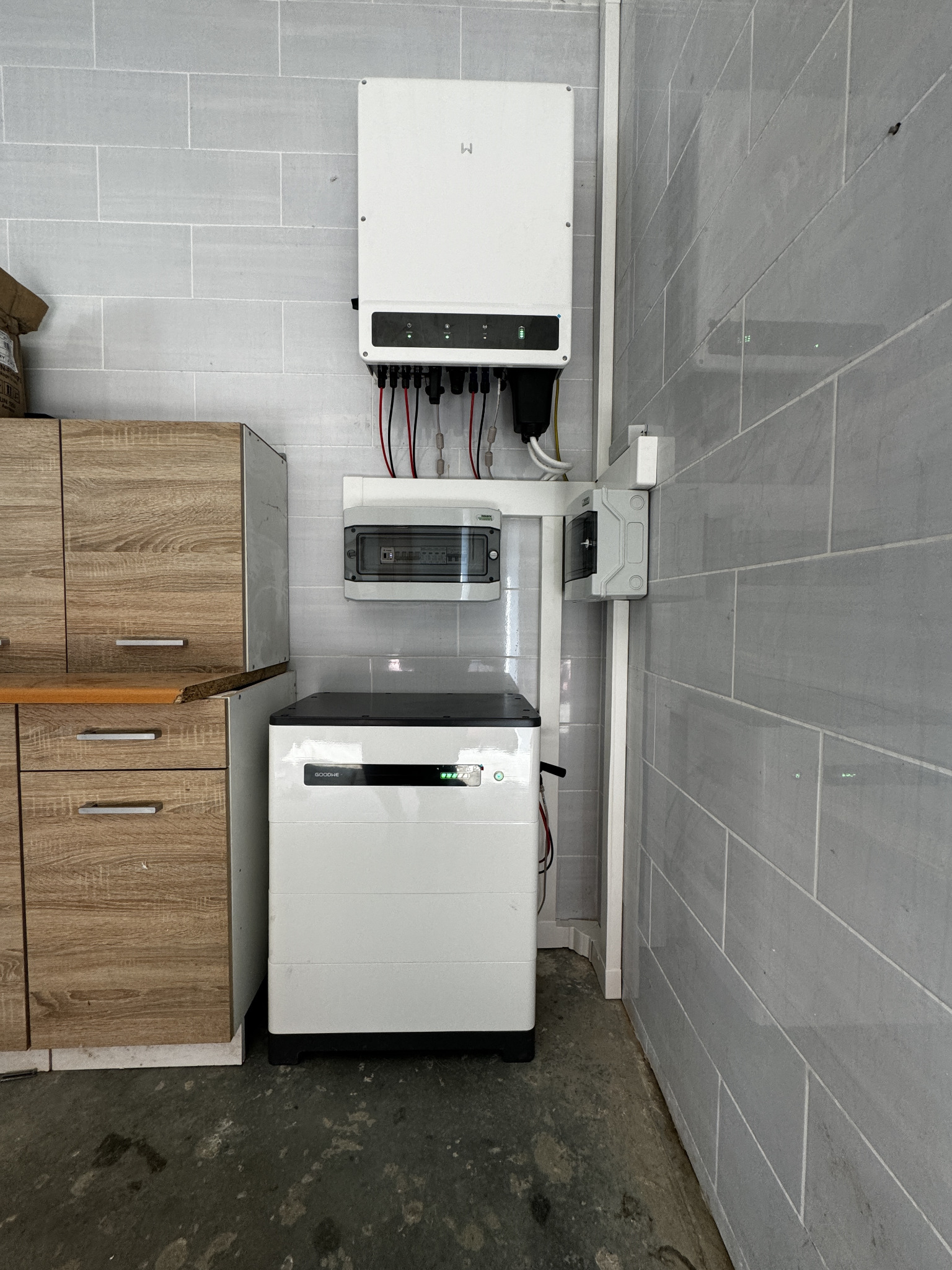 Biały magazyn energii Goodwe obok szafki w pomieszczeniu z szarymi płytkami, widoczny inwerter i skrzynka elektryczna.
