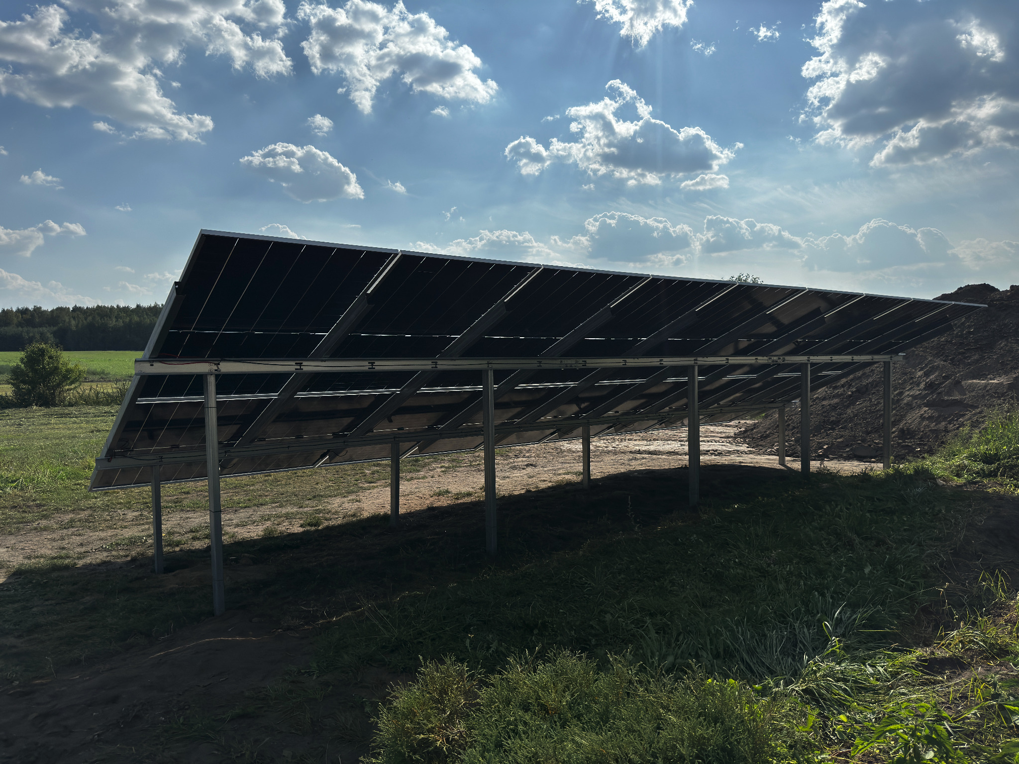 Widok od spodu na panele słoneczne zamontowane na metalowej konstrukcji, z widocznym niebem z chmurami w tle, a także fragmentem pola z zielenią i niewielkim pagórkiem ziemi.
