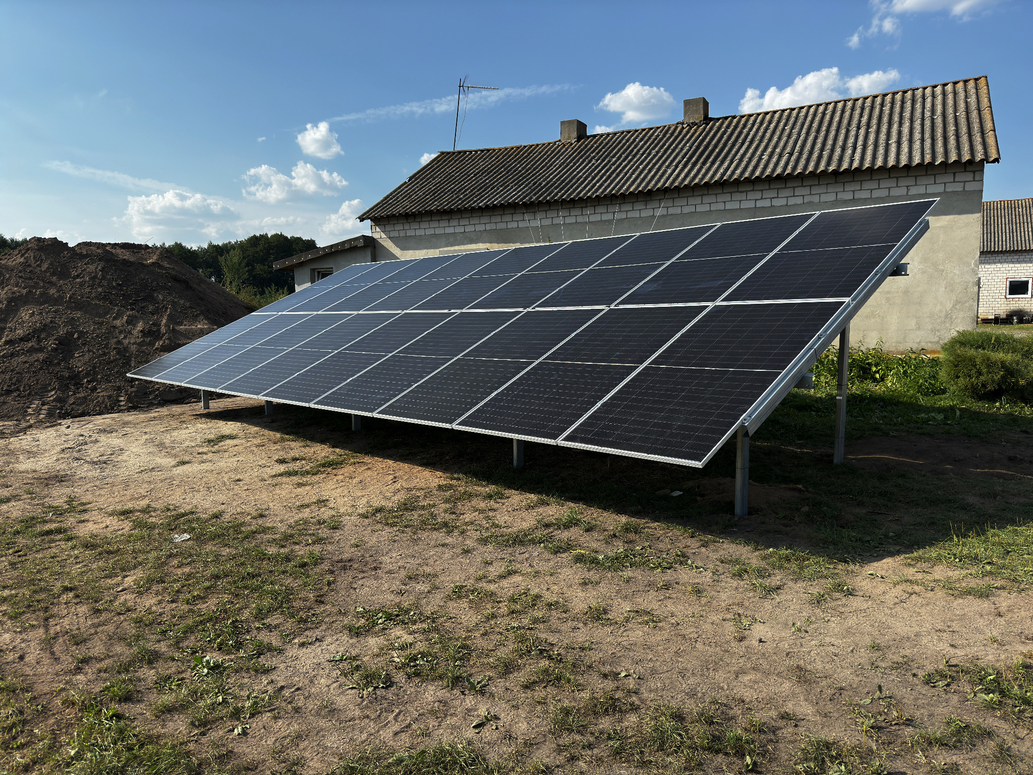 Instalacja paneli słonecznych na gruncie przed budynkiem z szarej cegły i stertą ziemi w tle, błękitne niebo z chmurami.