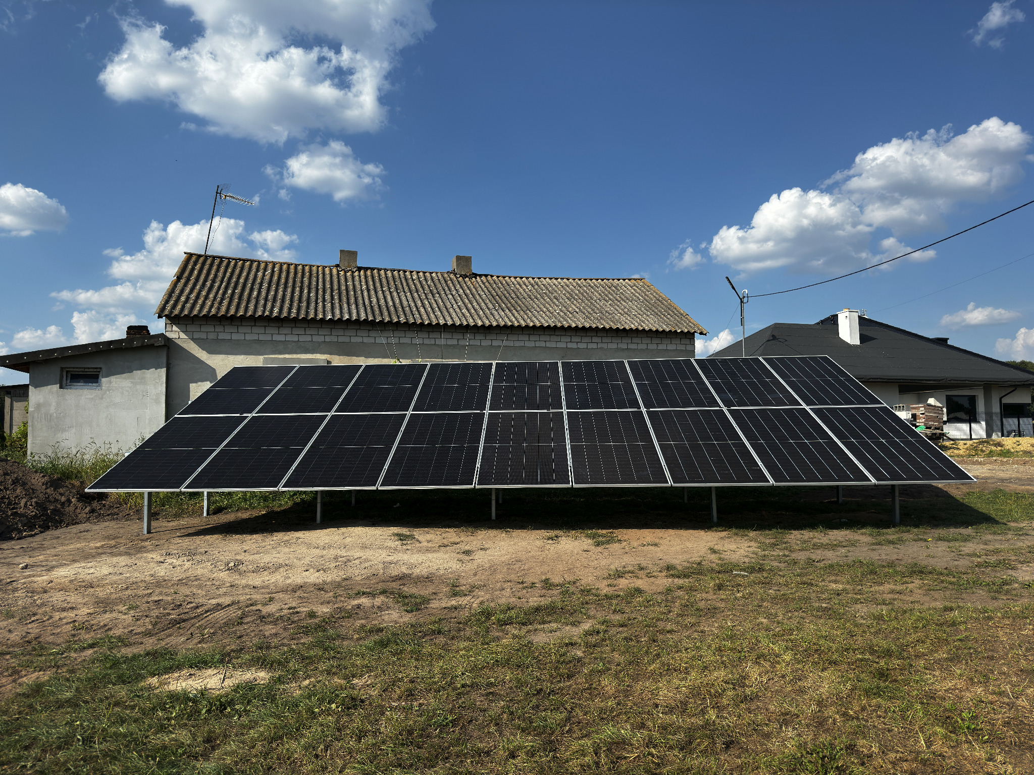 Naziemna instalacja fotowoltaiczna w trakcie montażu, widoczne panele na metalowej konstrukcji, w tle budynek z anteną i dom z ciemnym dachem, błękitne niebo z chmurami.
