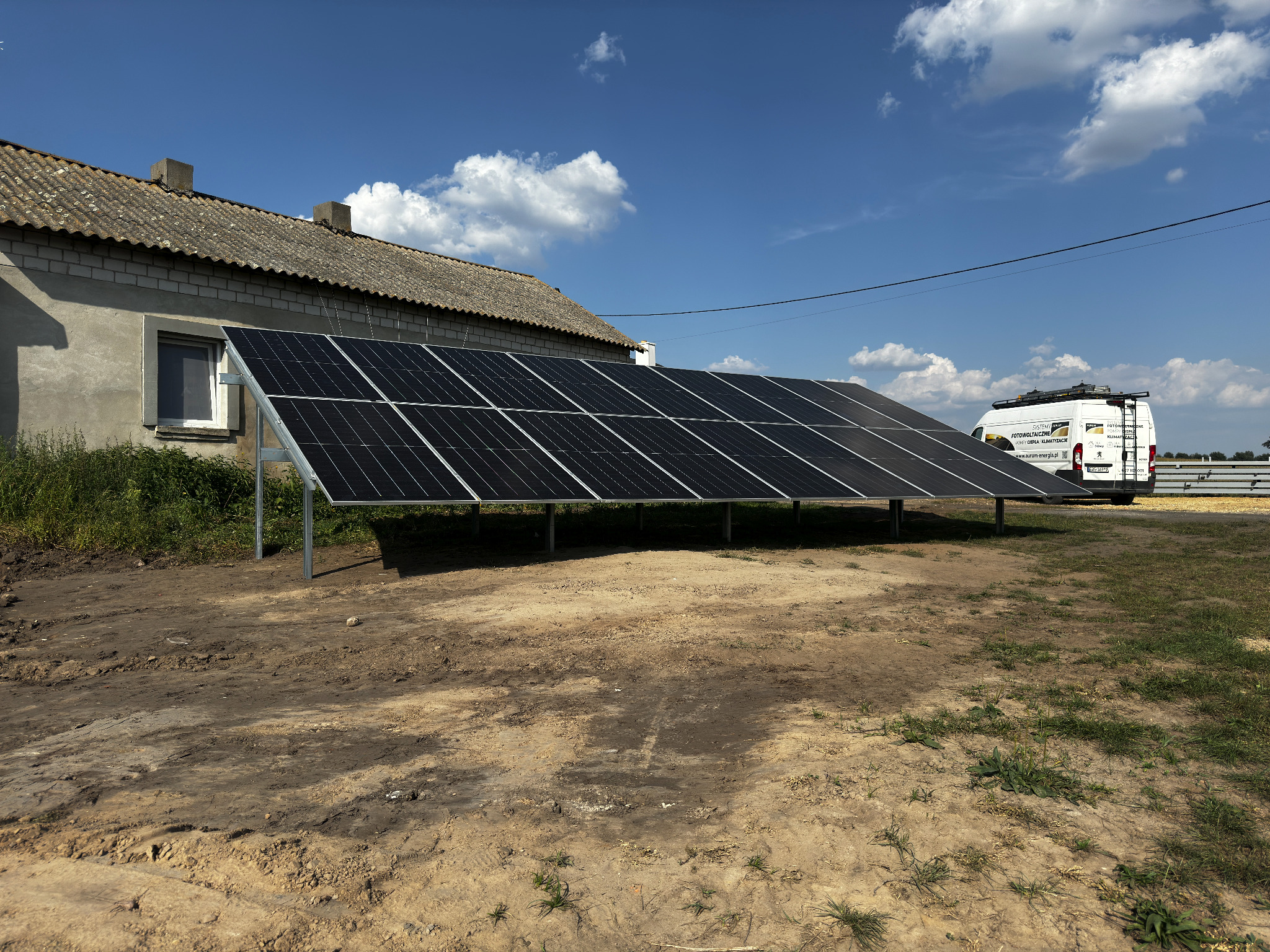 Panel słoneczny ustawiony na ziemi przed budynkiem mieszkalnym z zaparkowanym samochodem dostawczym w tle pod błękitnym niebem z chmurami.
