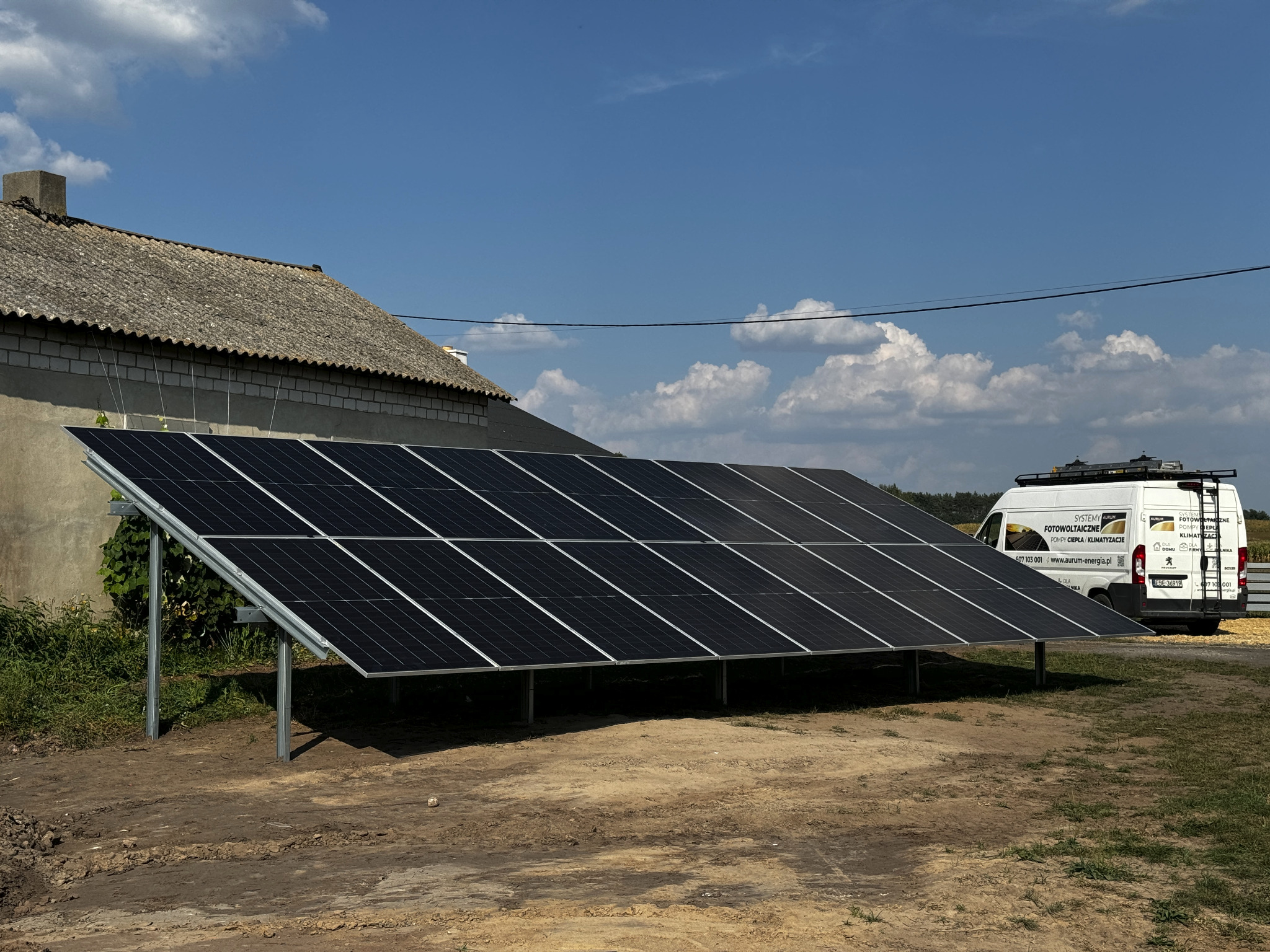Panele słoneczne zamontowane na konstrukcji na ziemi, obok budynku gospodarczego i samochodu dostawczego z reklamą firmy instalującej systemy fotowoltaiczne.