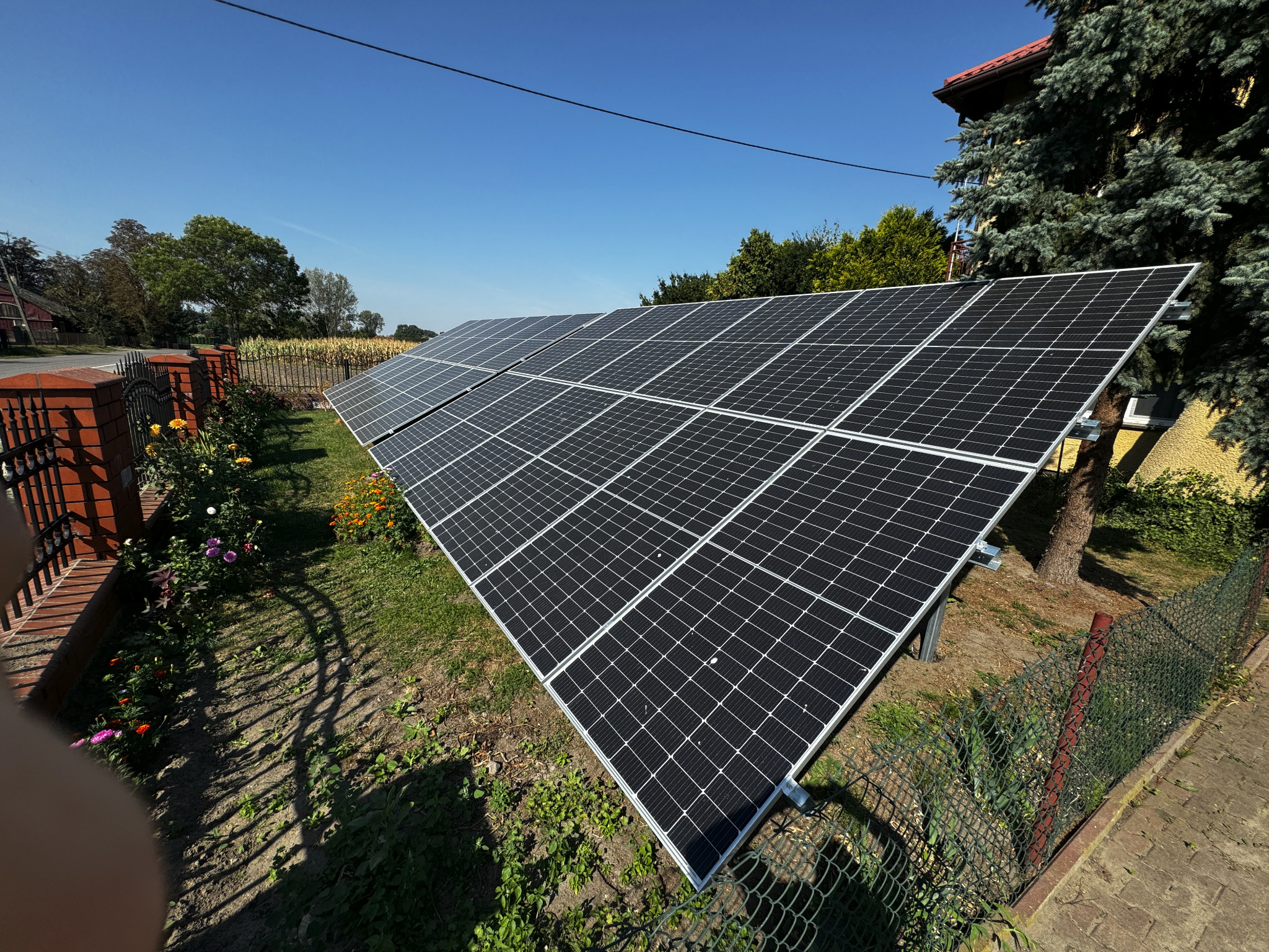 Instalacja fotowoltaiczna na gruncie w wiejskim otoczeniu, widoczna zza ozdobnego metalowego płotu i kwitnących kwiatów, na tle pola kukurydzy i błękitnego nieba.