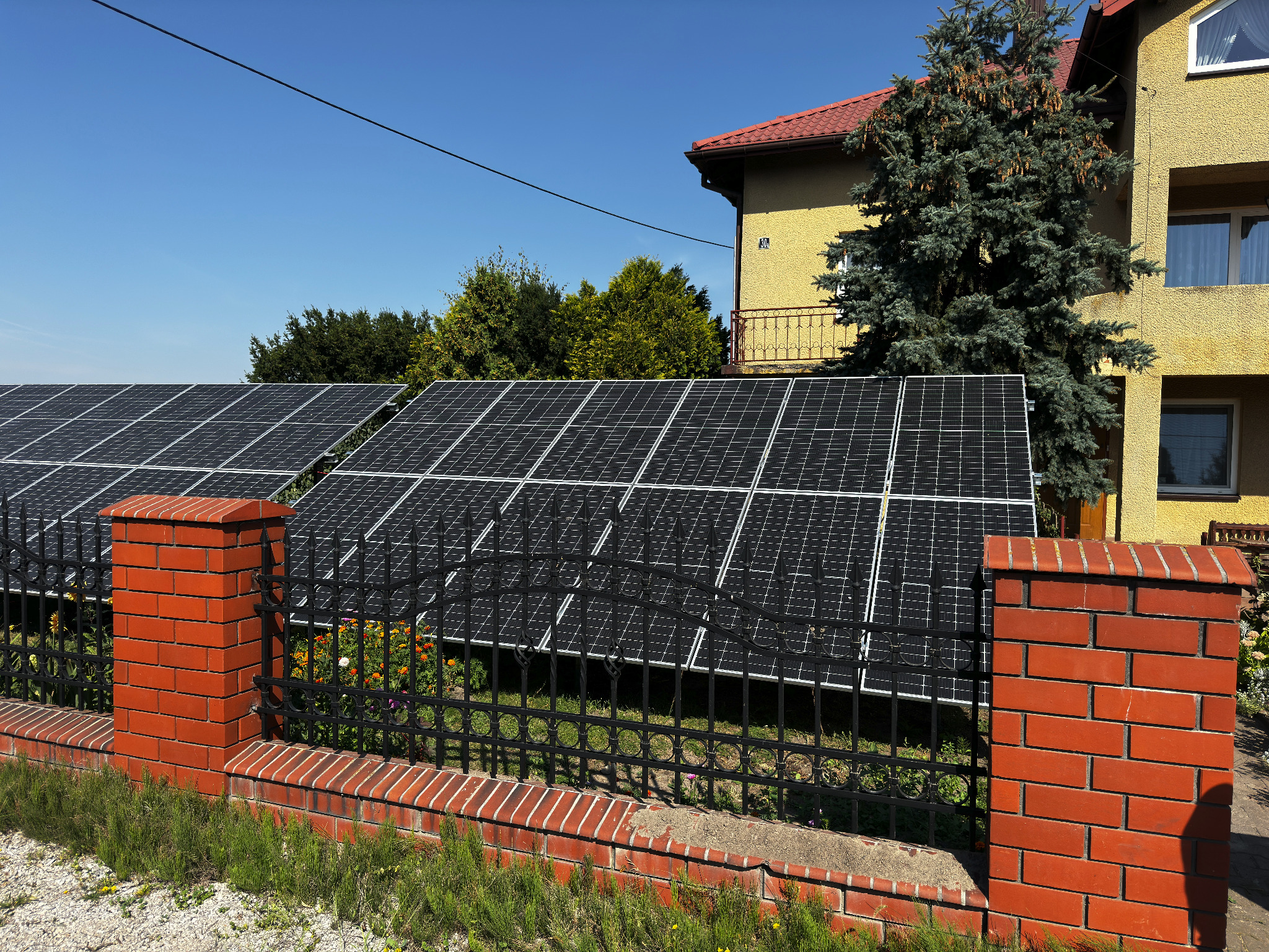 Instalacja paneli słonecznych za ozdobnym, niskim, ceglanym ogrodzeniem z czarnymi, metalowymi przęsłami, na tle żółtego domu z czerwoną dachówką i zielenią drzew