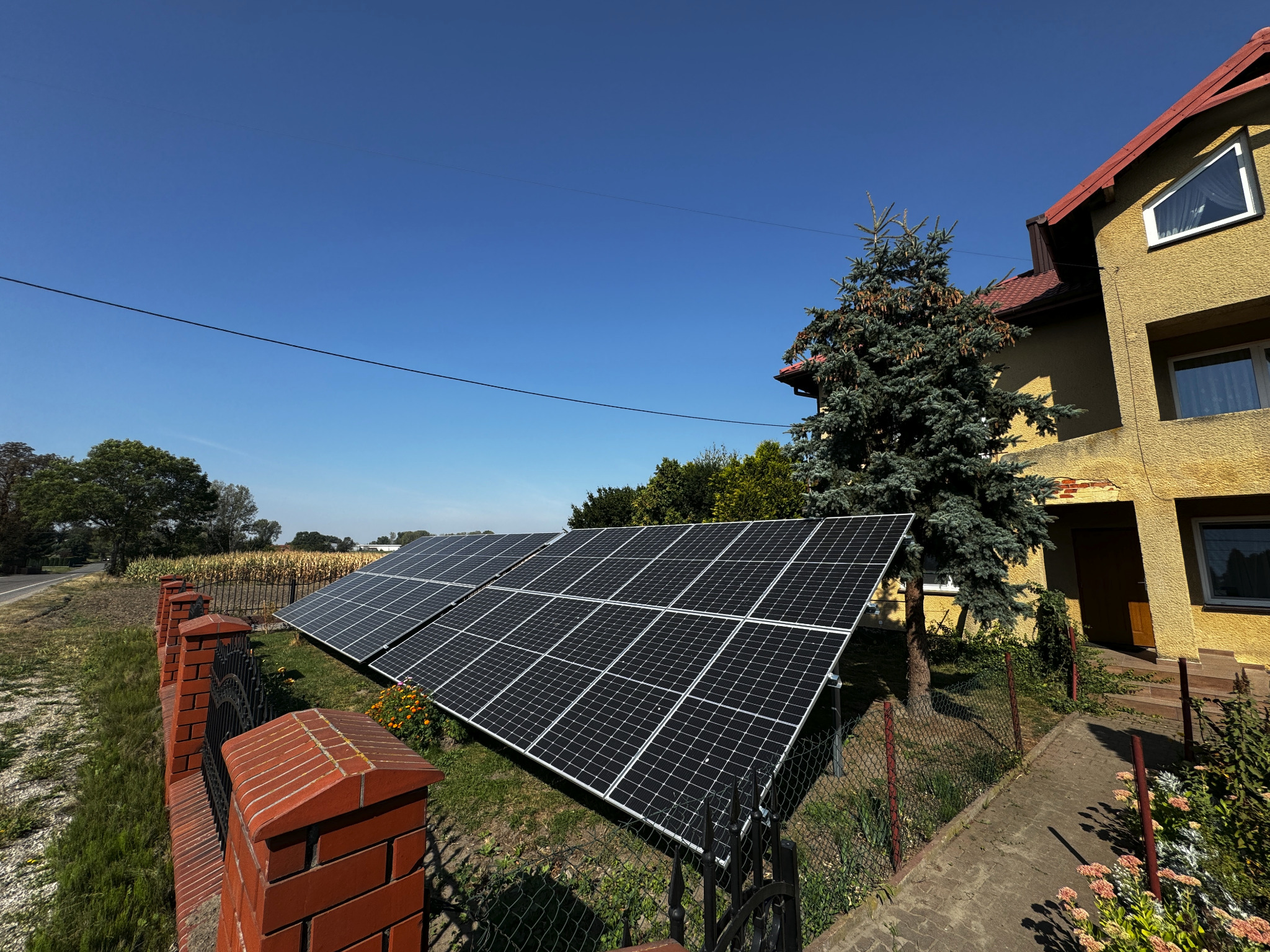 Panele słoneczne zamontowane na trawniku obok ceglanego ogrodzenia, z widokiem na dom z żółtą elewacją i świerkiem, w tle pole kukurydzy.