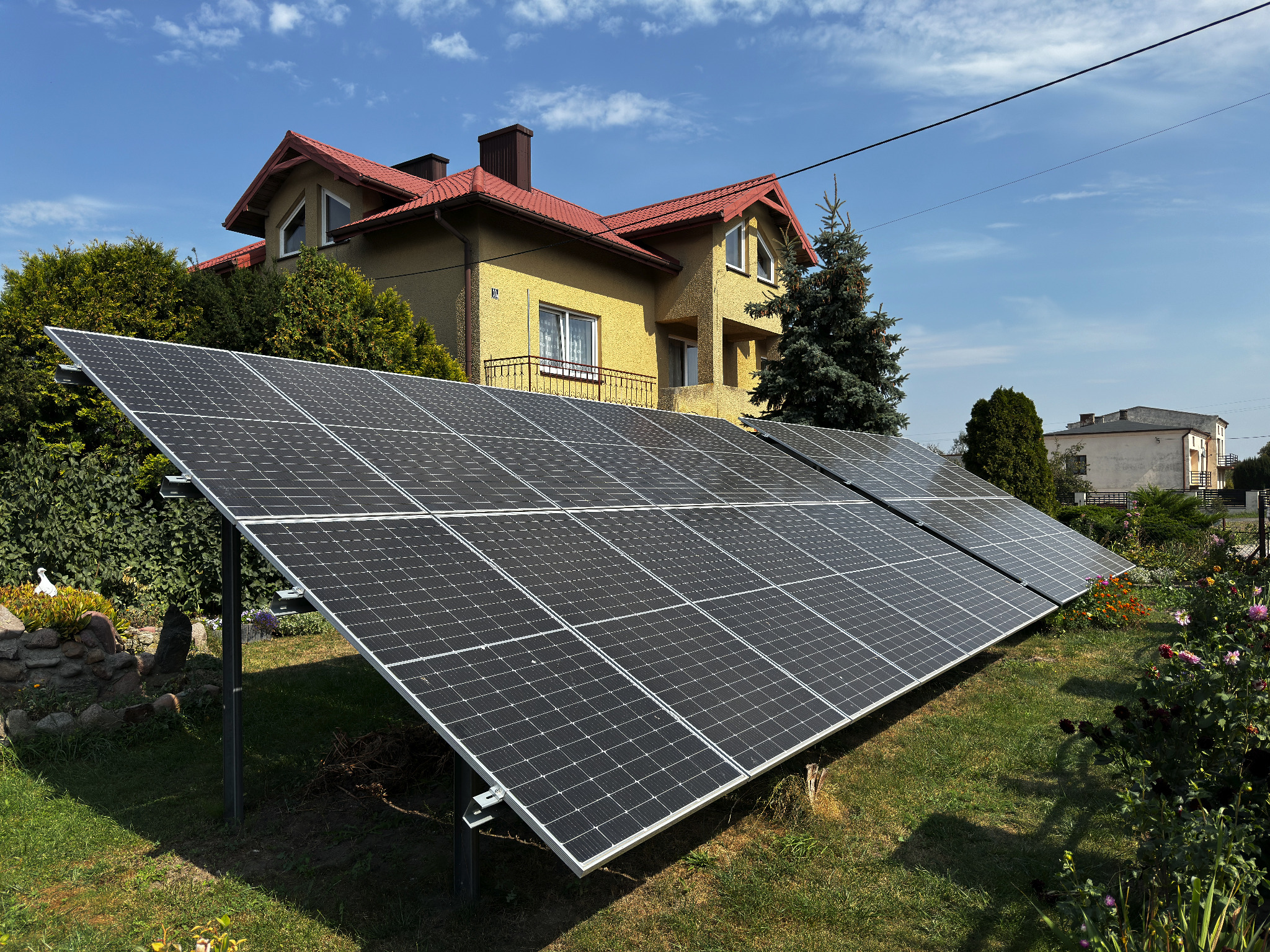 Panele fotowoltaiczne na trawniku przed domem z czerwoną dachówką, widok z niskiej perspektywy, słoneczny dzień.