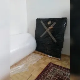 Obraz zabezpieczony folią stretch z naklejoną taśmą w kształcie litery X, obok rolka folii bąbelkowej i dywanik na parkiecie, gotowe do transportu.