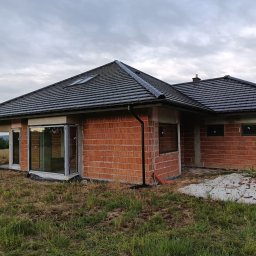 F.U.H.P HatBud Marcin Toboła - Dom w trakcie budowy z widocznymi oknami PCV, murowanymi ścianami i ciemną dachówką. Widok na zielony teren i pochmurne niebo w tle.