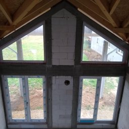 F.U.H.P HatBud Marcin Toboła - Montaż okien PCV w surowym stanie domu z widoczną konstrukcją dachu i bloczkami. Dwa okna trójkątne na górze i dwa prostokątne na dole. Widok na zewnątrz.