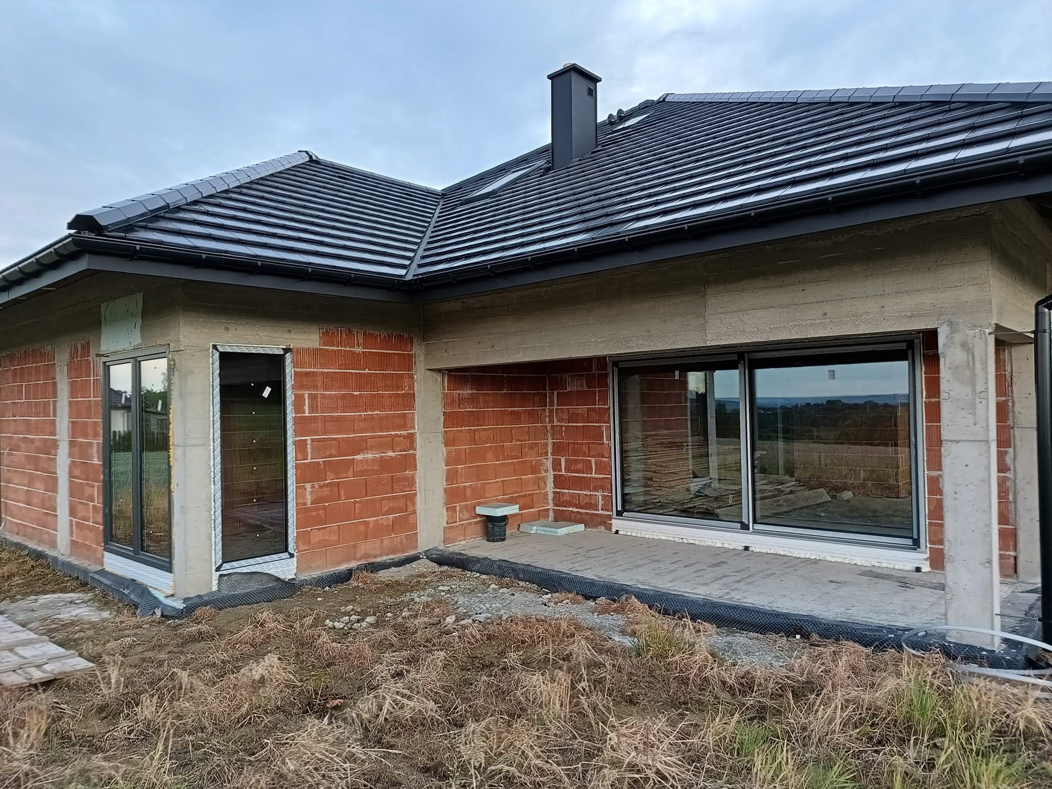 Surowy stan domu z ciemną dachówką, widoczne okna z czarnymi ramami i niedokończona elewacja z cegły. Fragment terenu z roślinnością.