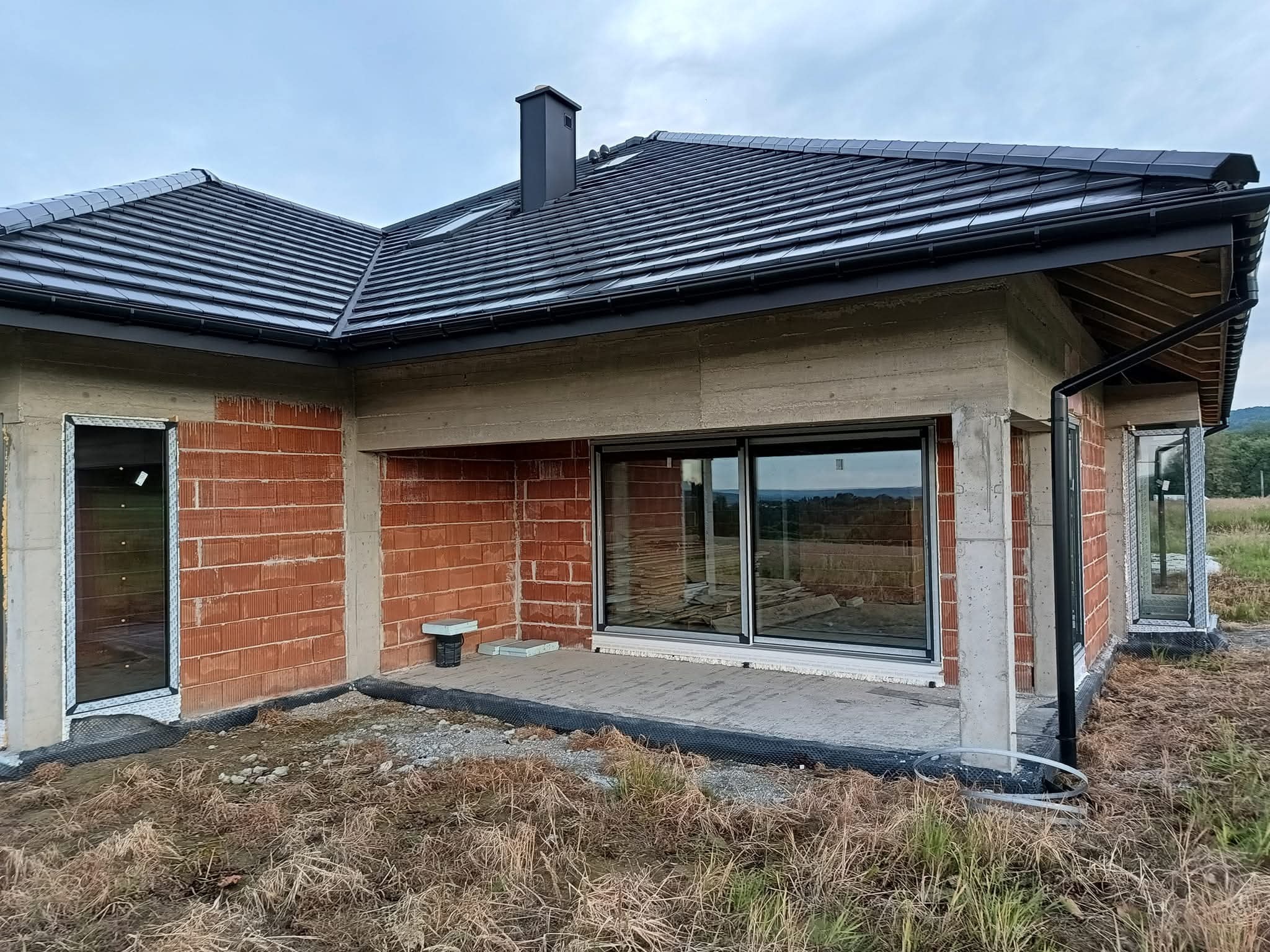 Surowy stan domu jednorodzinnego z czarną dachówką i oknami z czarnymi ramami, widok z zewnątrz. Budynek w trakcie budowy, widoczne niedokończone ściany.