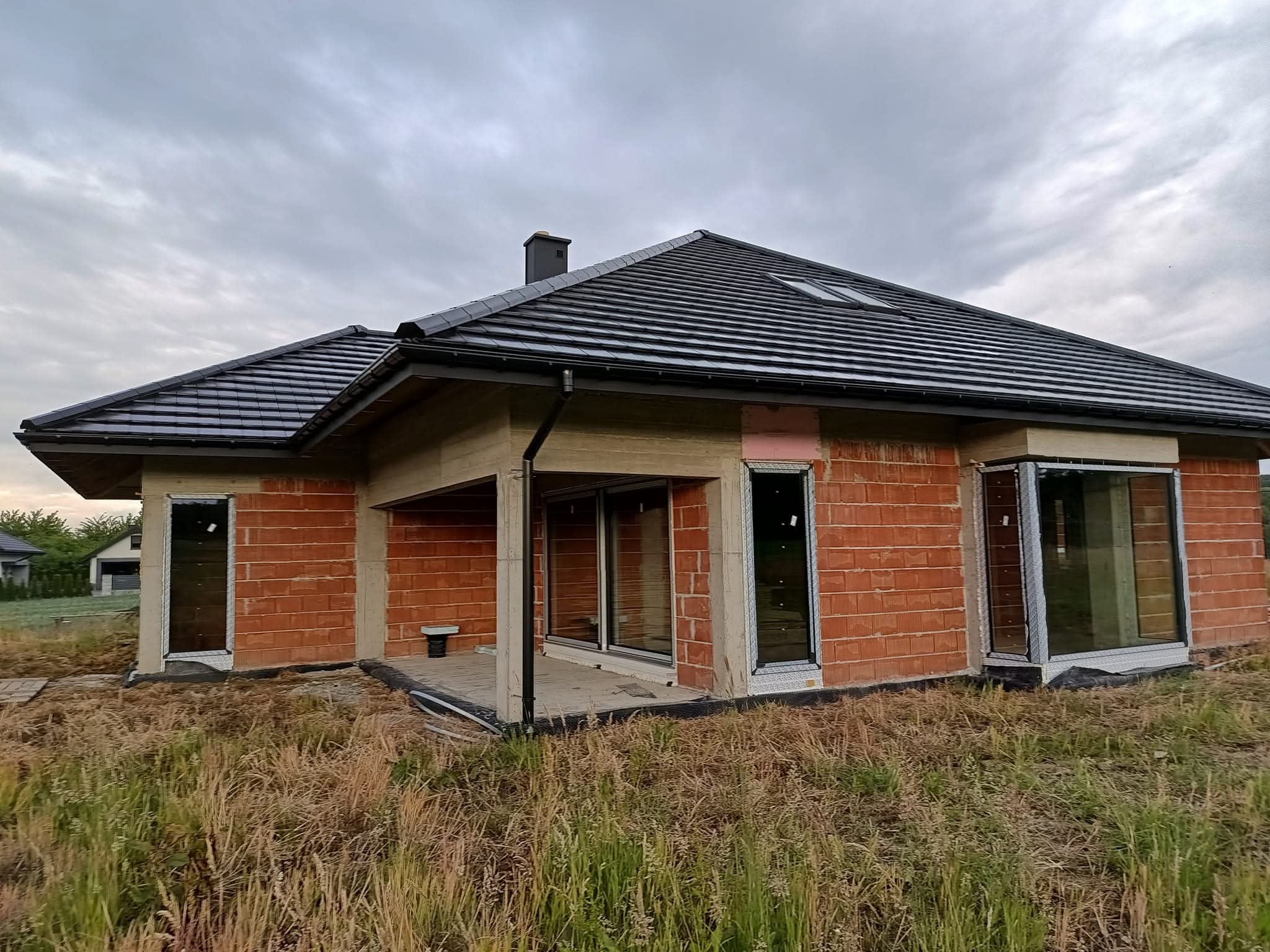 Dom jednorodzinny w trakcie budowy, widoczne okna, czarna dachówka na dachu i czerwona cegła na ścianach. Pochmurne niebo w tle. Widok z poziomu gruntu.